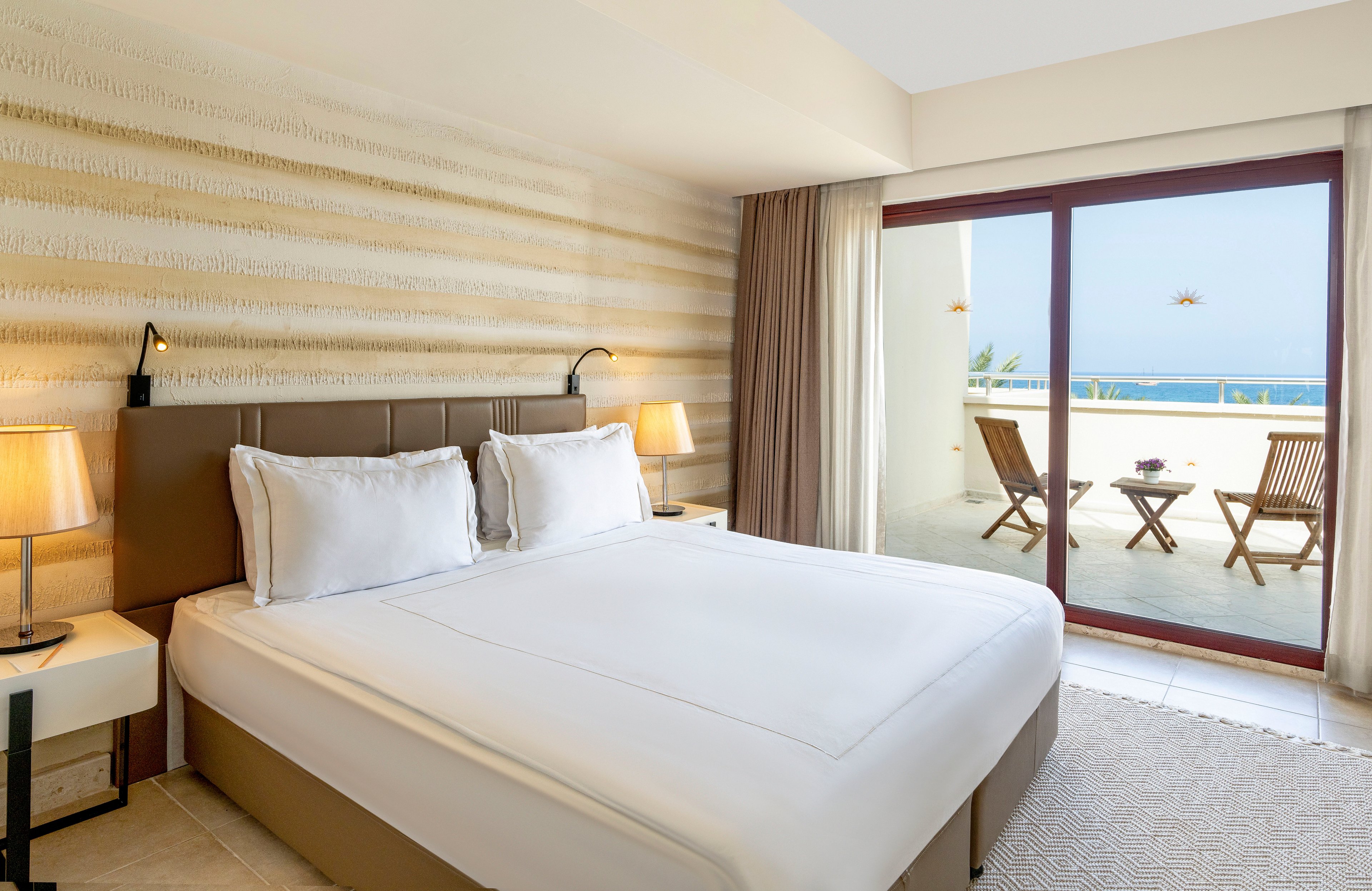 Modern hotel bedroom with a large neatly made bed and soft neutral decor. Two bedside tables with warm lamps flank the bed, creating a cozy and inviting atmosphere. A sliding glass door opens to a balcony with wooden chairs and a small table overlooking the sea. Natural light floods the room, highlighting the clean, minimalist style and coastal setting.