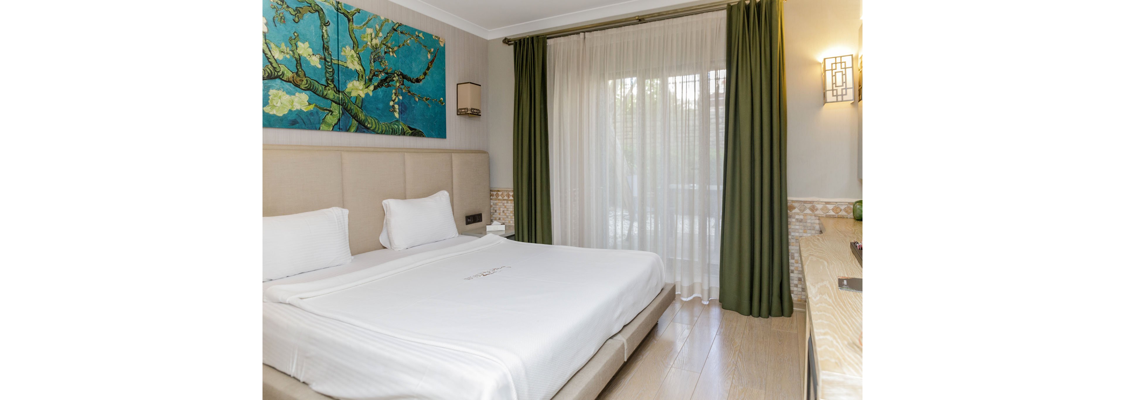 A neatly made double bed with white linens sits in a modern hotel room. Above the upholstered headboard hangs a large colorful painting of flowering branches. Floor-to-ceiling windows are covered with sheer white curtains and dark green drapes, letting in soft natural light. The room features light wood flooring, wall sconces, and a neutral color palette with green accents for a calm, contemporary feel.