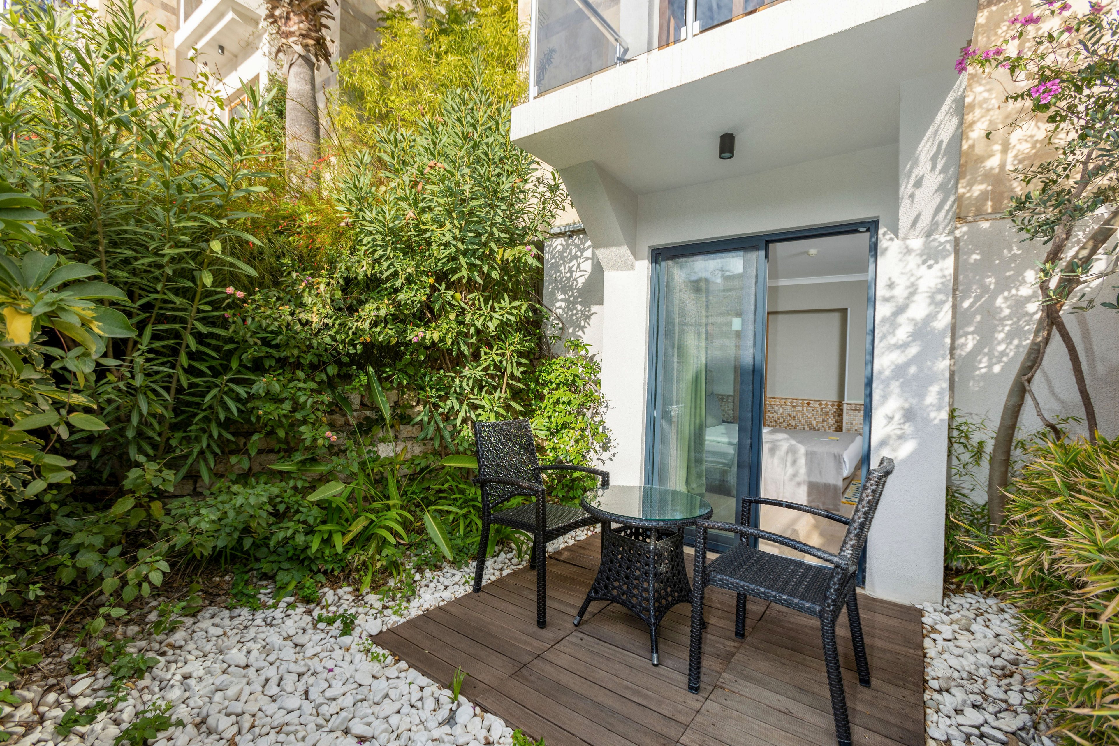 A small outdoor patio features a round glass-top table with three black wicker chairs on a wooden deck. The space is surrounded by lush green plants and white decorative stones, creating a private garden feel. A large sliding glass door opens into a bright interior room, allowing natural light to flow between indoors and outdoors. The scene suggests a relaxing spot for conversation, reading, or enjoying a drink in a modern residential setting.