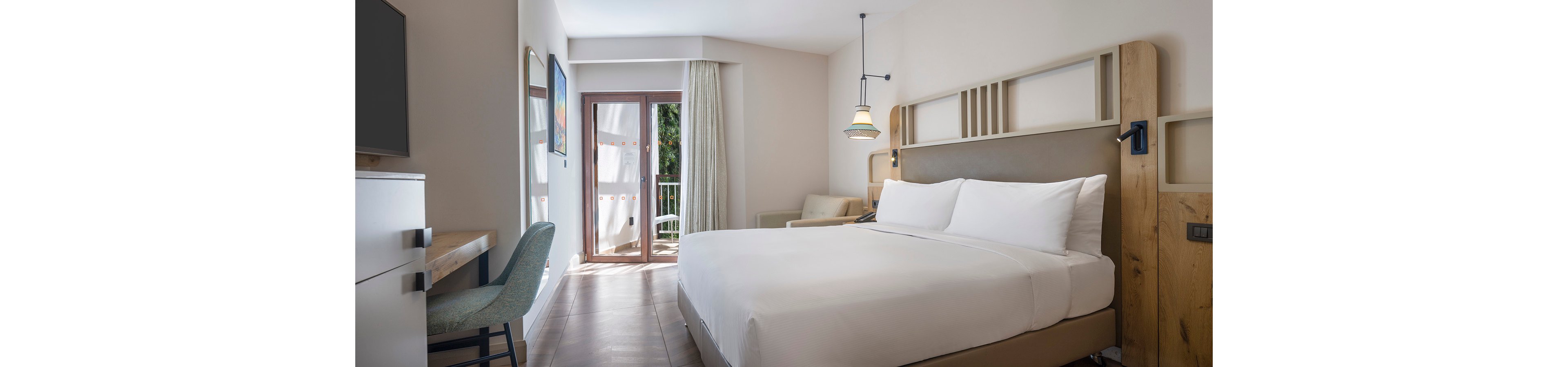 A contemporary hotel room features a large neatly made bed with white linens and wooden headboard. Natural light streams in through glass doors leading to a balcony with greenery outside. The room includes a desk with a chair, a sofa, and minimalist decor in soft neutral tones. Subtle artwork and pendant lighting add a relaxed, welcoming atmosphere.