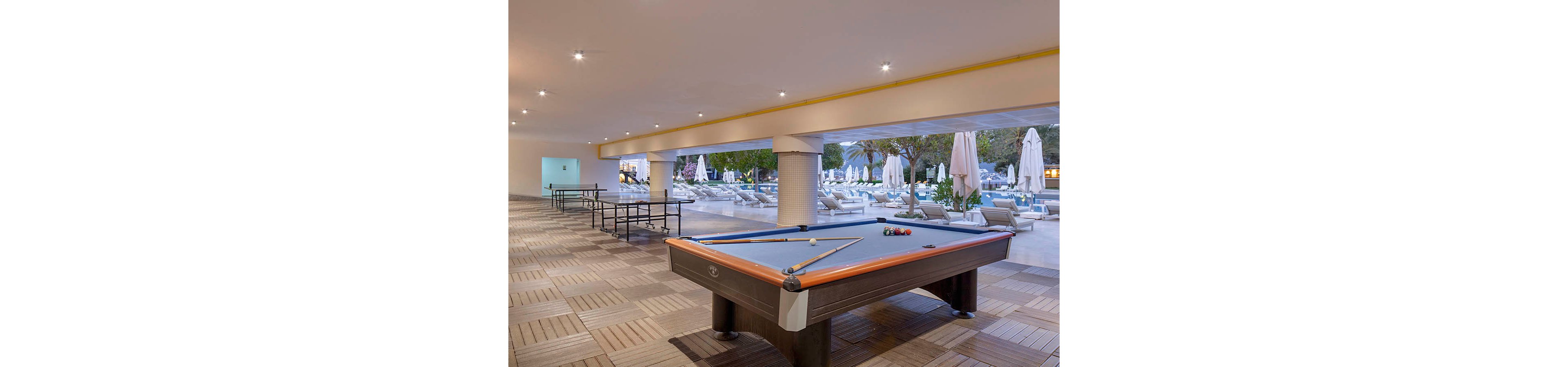 An indoor billiards table with balls and cues is set in a spacious covered recreation area. Additional game tables line the tiled floor, leading the eye toward an outdoor pool deck. Beyond the open wall, sun loungers and umbrellas surround the swimming pool amid trees and greenery. Soft ceiling lighting and neutral tones create a relaxed vacation atmosphere suitable for leisure and socializing.