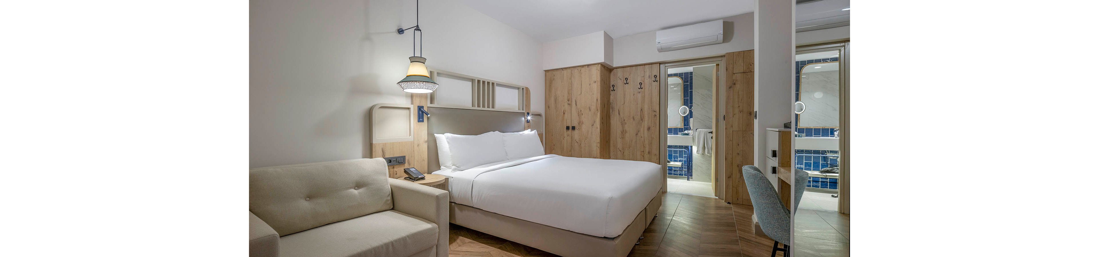 Contemporary hotel room featuring a large double bed with white linens and a light-colored sofa. Wooden wardrobes and cabinetry add warmth to the minimalist interior design. A desk and chair sit near the entrance to the tiled ensuite bathroom, visible through glass doors. Soft lighting and neutral tones create a calm, inviting atmosphere suitable for guests.