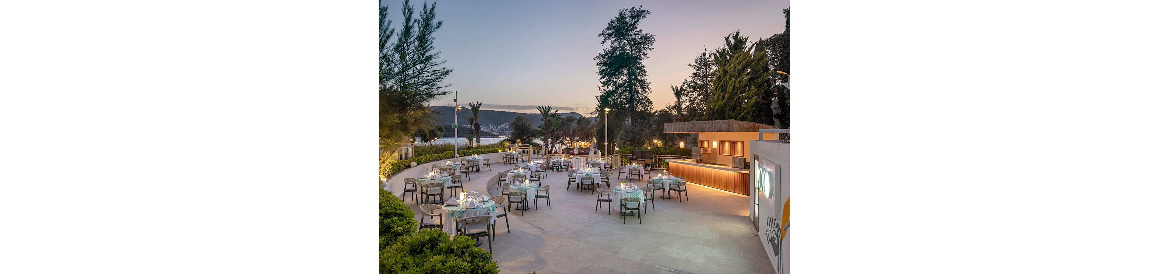 An elegant open-air restaurant terrace is set with multiple tables overlooking a calm waterfront and distant hills at dusk. Tables are arranged across a spacious stone patio, each prepared with glassware, plates, and napkins for evening dining. Warm lighting from the bar area and subtle ground lights contrasts with the cool twilight sky. Lush trees and greenery frame the scene, creating a relaxed, upscale coastal atmosphere.