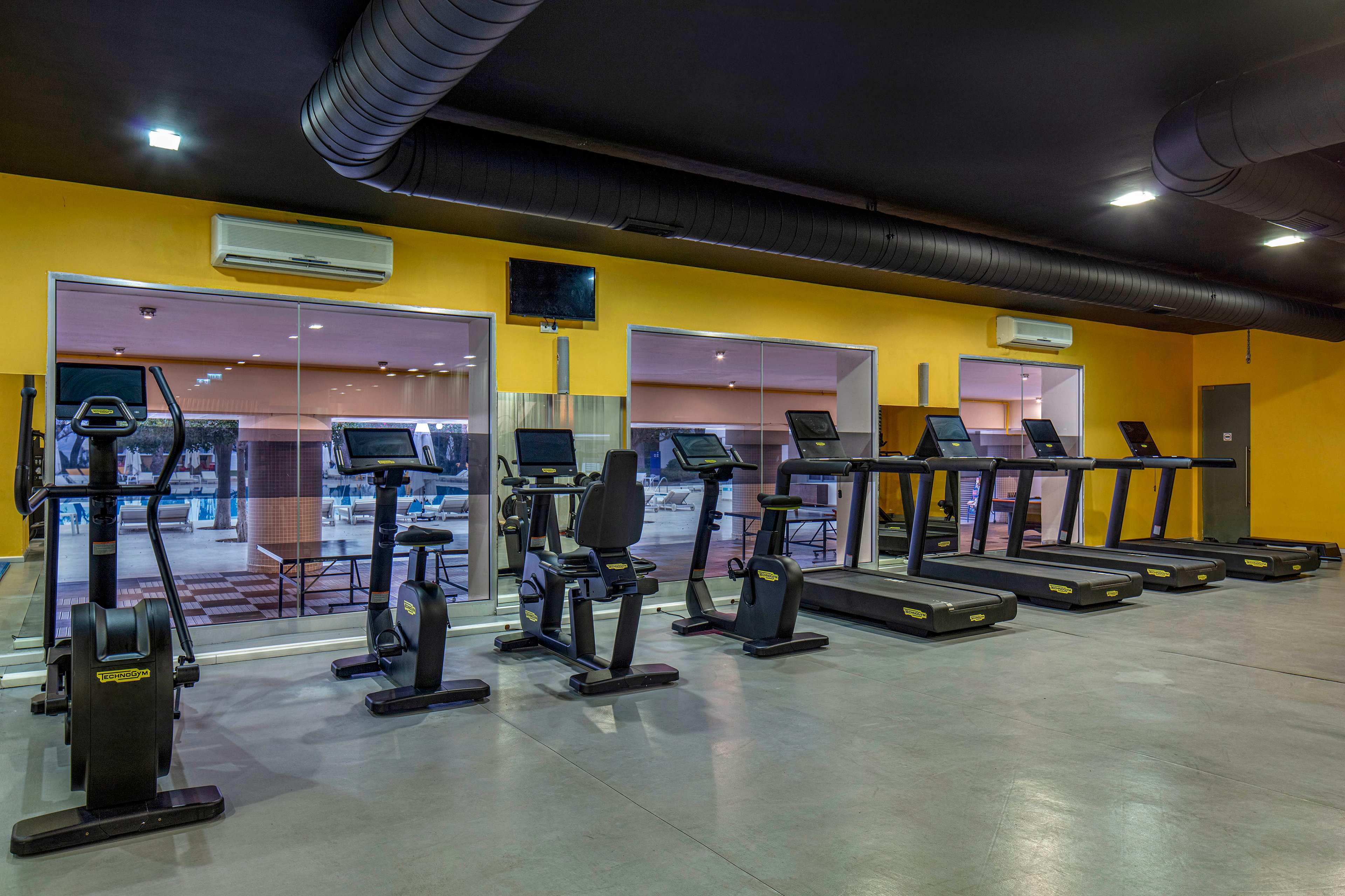 A row of treadmills and stationary bikes is arranged neatly in an empty, modern gym. The room features bright yellow walls, large mirrors, and glass windows looking out onto another area of the facility. Overhead air conditioning units and exposed ductwork contribute to the contemporary industrial style. The clean, polished floor and organized machines suggest a professional fitness environment ready for use.