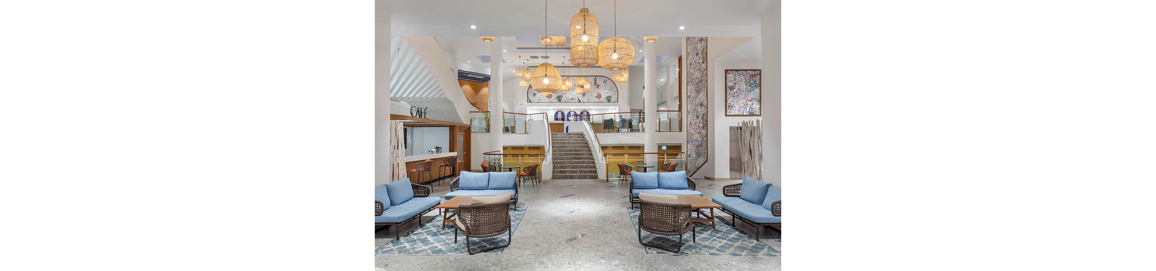 Spacious contemporary hotel lobby featuring blue cushioned lounge seating and woven armchairs arranged around low tables. A central staircase leads to an upper level, framed by white columns and decorative railings. Warm pendant lights with wicker shades hang from the ceiling, complementing patterned rugs and abstract wall art. A café area is visible to the left, adding to the welcoming, social atmosphere of the space.