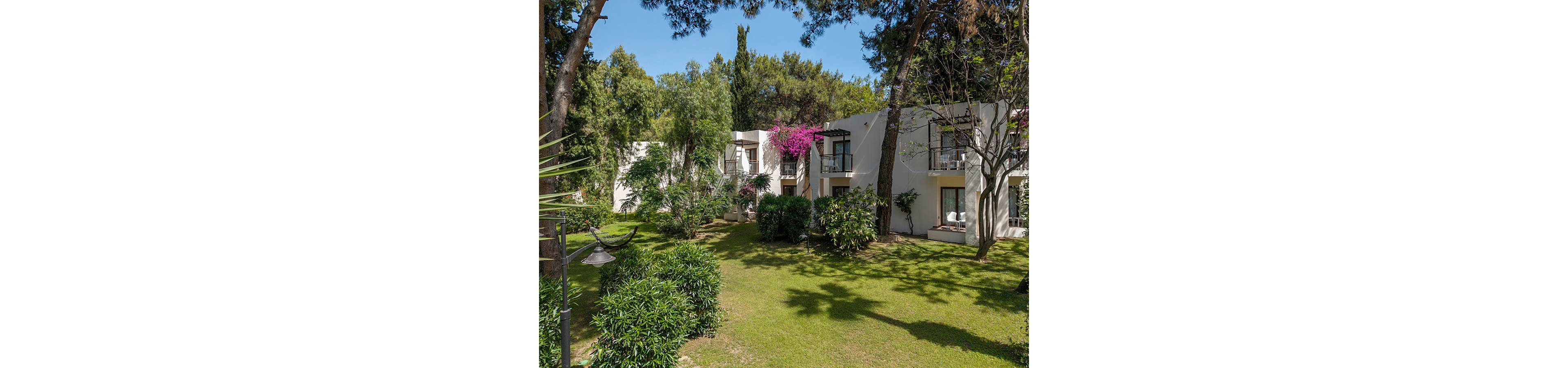 The image shows a low-rise modern apartment or hotel building nestled within a landscaped garden. White exterior walls and small balconies are partially covered by vibrant pink flowering vines. Tall trees and dense shrubs frame the building, casting soft shadows across the well-maintained green lawn. The overall scene suggests a peaceful, sunny retreat in a natural setting.