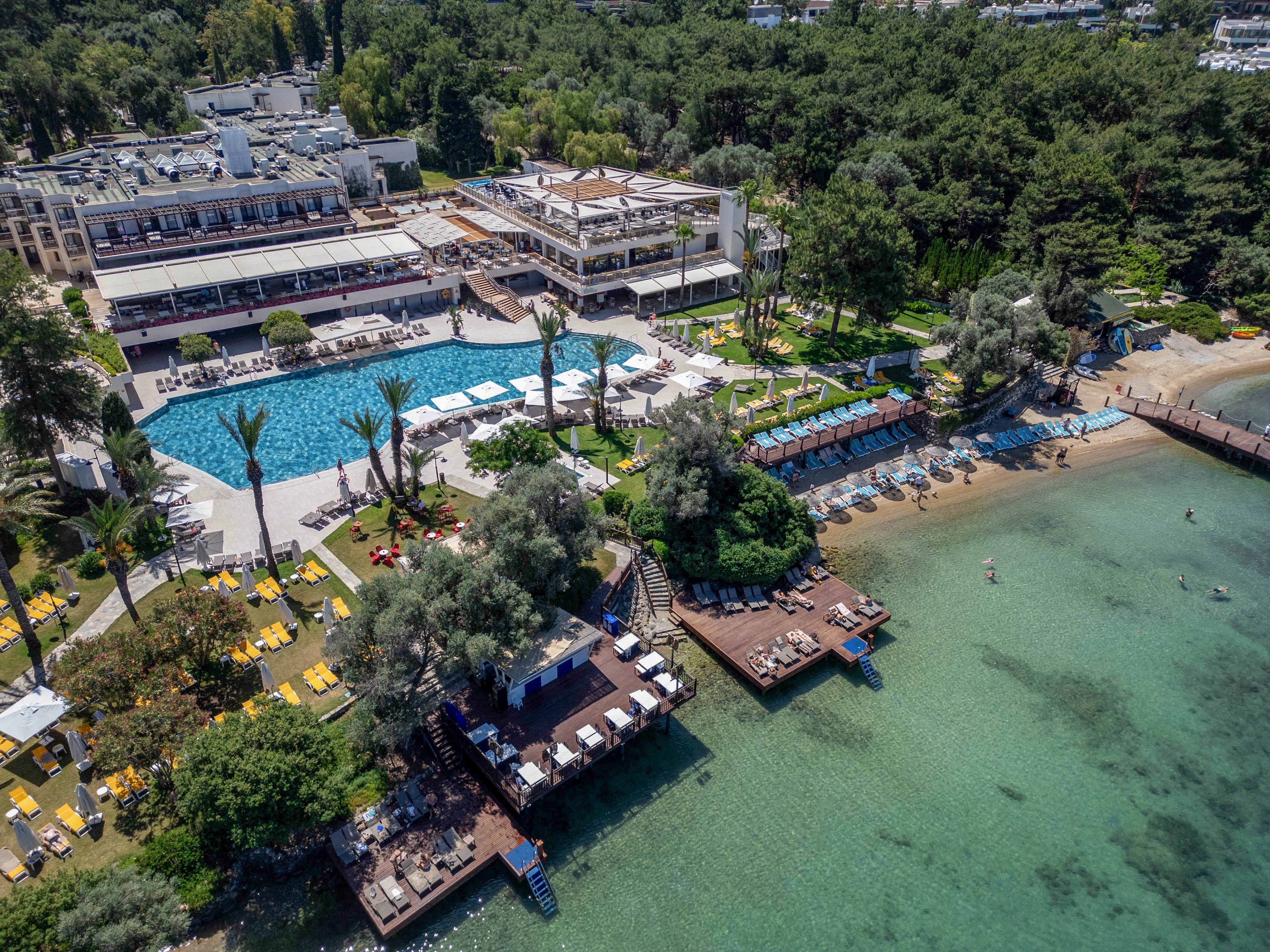 A large coastal resort complex is captured from above, featuring a central outdoor swimming pool surrounded by sun loungers and umbrellas. Multiple wooden decks extend over the clear turquoise water, providing additional seating areas along the shoreline. The sandy beach and shallow sea host a few people enjoying the water, while dense green trees frame the property in the background. The scene conveys a relaxed vacation atmosphere with a mix of built structures and natural landscape.
