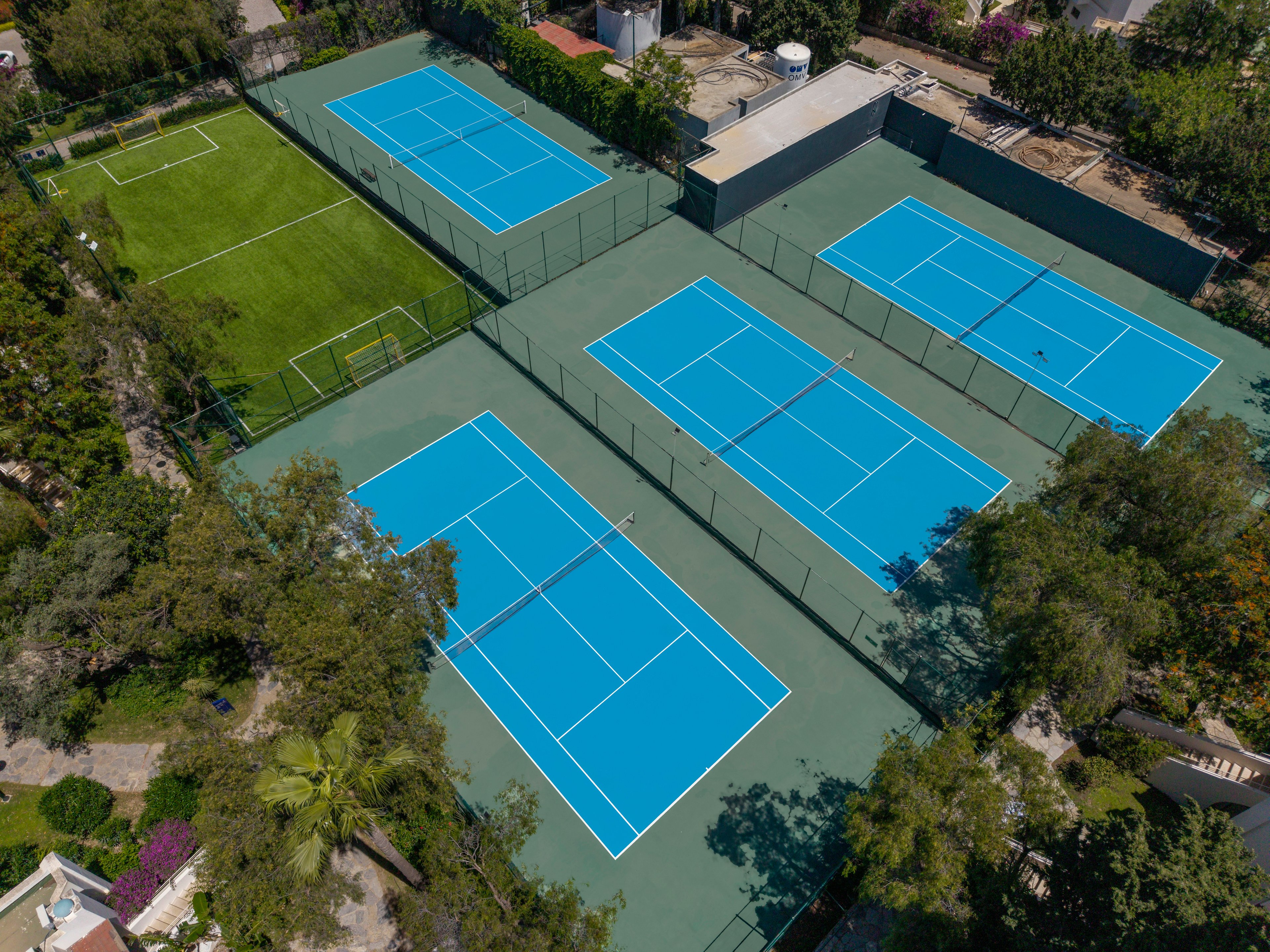Aerial view of a sports complex featuring several bright blue tennis courts surrounded by trees. The courts are arranged side by side on a green surface, enclosed by high fencing. Adjacent to the tennis area is a rectangular grass field with soccer goals, adding variety to the recreational space. The scene is captured on a clear day, highlighting the vivid colors and orderly layout of the facilities.