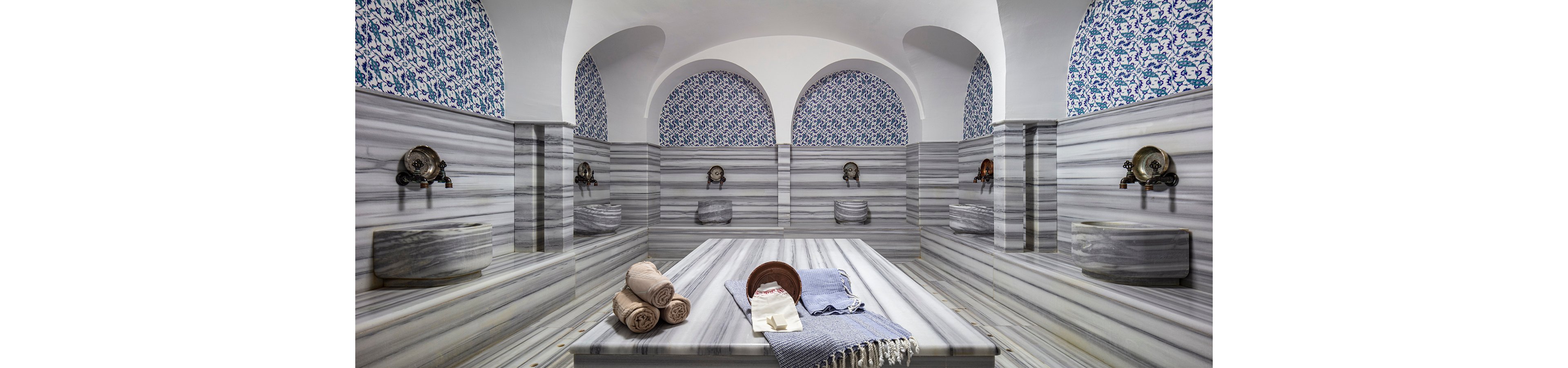 Spacious marble-lined Turkish hammam with a central massage platform and folded towels arranged neatly on top. Ornate basins and metal taps line the walls beneath arched niches, creating a symmetrical and elegant layout. Blue and white patterned tiles decorate the upper walls, adding a rich cultural detail to the serene bathing space. Soft, even lighting enhances the clean, tranquil atmosphere of the spa-like interior.