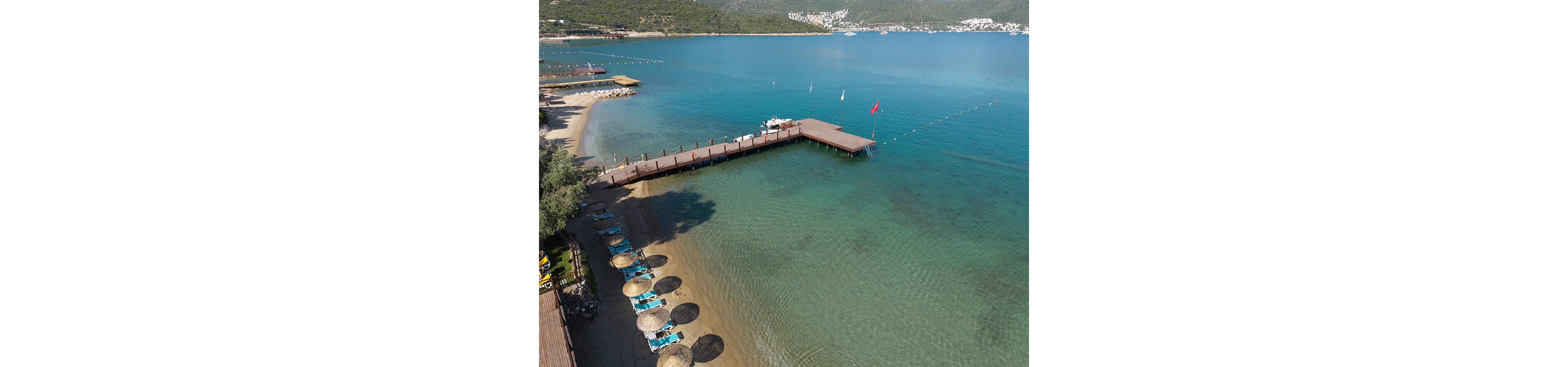 A wooden pier extends over calm, clear turquoise sea beside a sandy beach. Straw parasols and sun loungers line the shore, creating a relaxed holiday atmosphere. A red flag flies at the end of the pier, and floating buoys mark a swimming area in the water. Green hills and scattered white buildings are visible in the distant background across the bay.