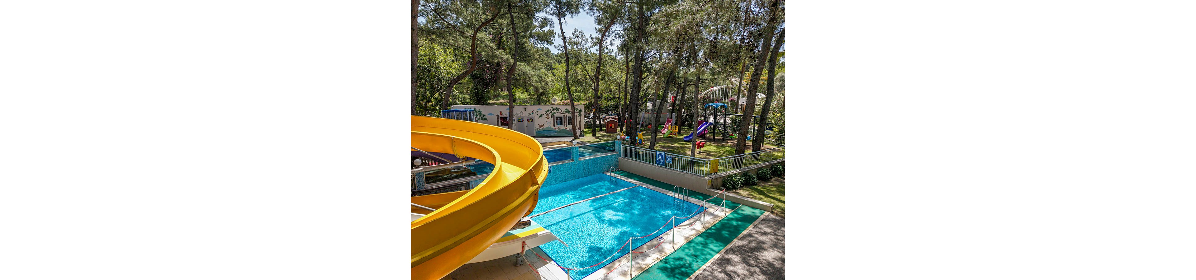 An outdoor swimming pool is shown with a large yellow spiral waterslide leading into the clear blue water. The pool area is surrounded by tall trees, creating a shaded forest-like setting. In the background, a fenced playground with colorful slides and play structures is visible on the grass. The scene suggests a family-friendly recreation area in a resort or park.