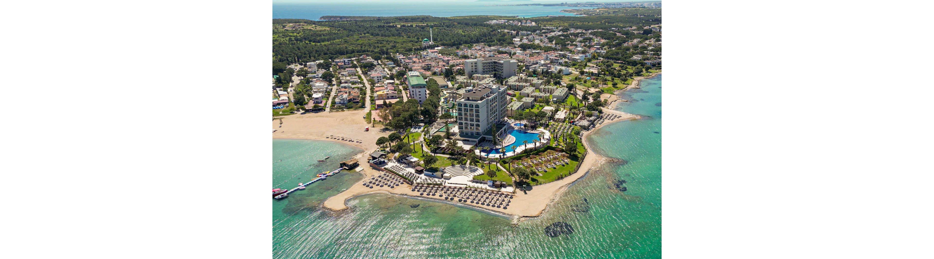 A large beachfront resort complex extends on a small peninsula surrounded by turquoise sea. The property features a tall hotel building, a large outdoor pool, and landscaped gardens with palm trees. Sun loungers and umbrellas line the sandy beach, and several boats are anchored near the shore. In the background, a coastal town and green forested areas stretch toward the horizon under clear skies.