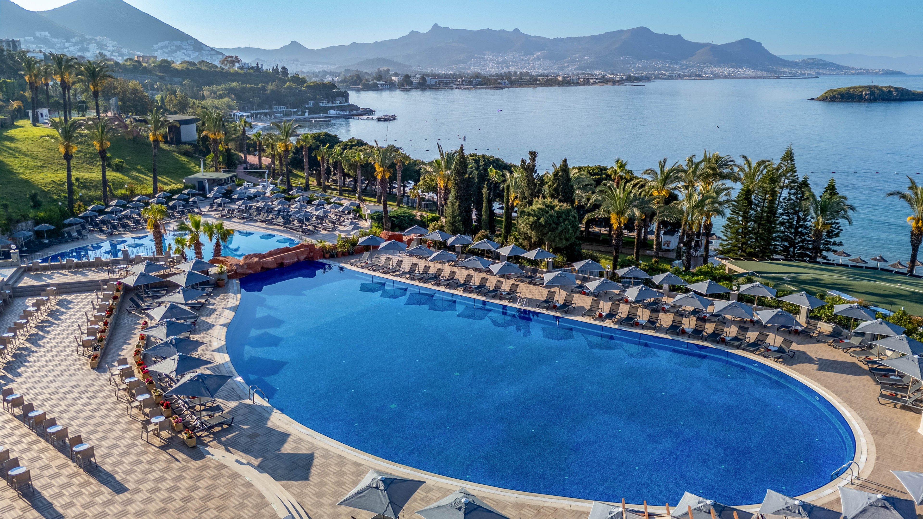 A large, curved swimming pool is the centerpiece of a luxury resort, surrounded by numerous lounge chairs and umbrellas. The setting features lush palm trees and manicured gardens, with a stunning view of a calm bay and distant mountains under a clear blue sky. The tranquil atmosphere and vibrant blue palette evoke a relaxing vacation mood. No visible text or numbers are present in the image.