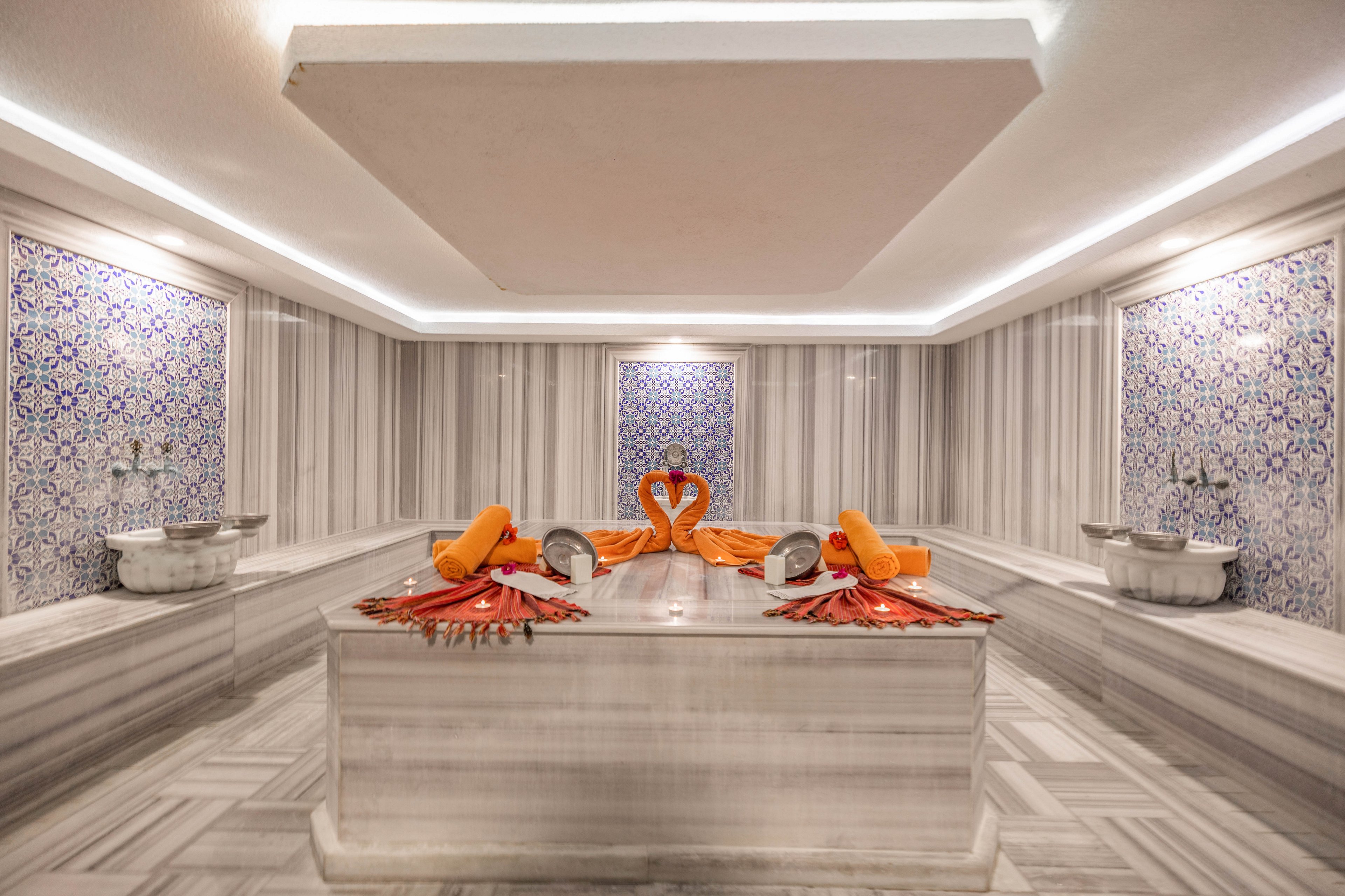 The image features a traditional Turkish hammam with a central marble platform, ornate blue tile walls, and two decorative towel arrangements shaped like swans. Orange towels and metal bowls are placed on the marble, adding vibrant color to the serene, spa-like setting. The space is well-lit, emphasizing the clean lines and relaxing ambiance.