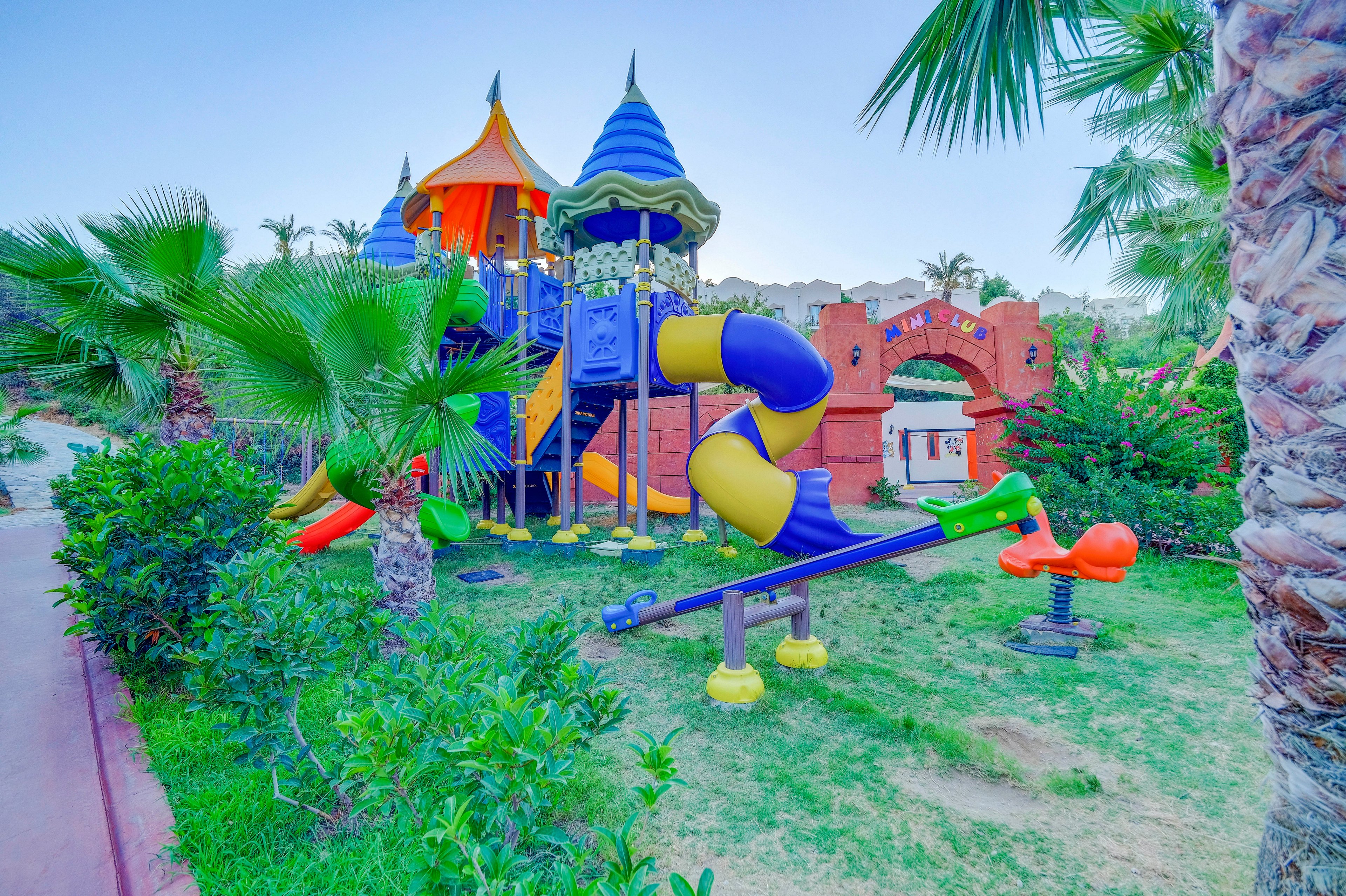 A vibrant children's playground features a large multi-level play structure with blue and yellow spiral slides and pointed roofs. The setting is an outdoor park surrounded by palm trees and lush greenery, with a seesaw and spring rider in the foreground. The background includes a decorative brick archway and clear blue sky, creating a cheerful and inviting atmosphere.