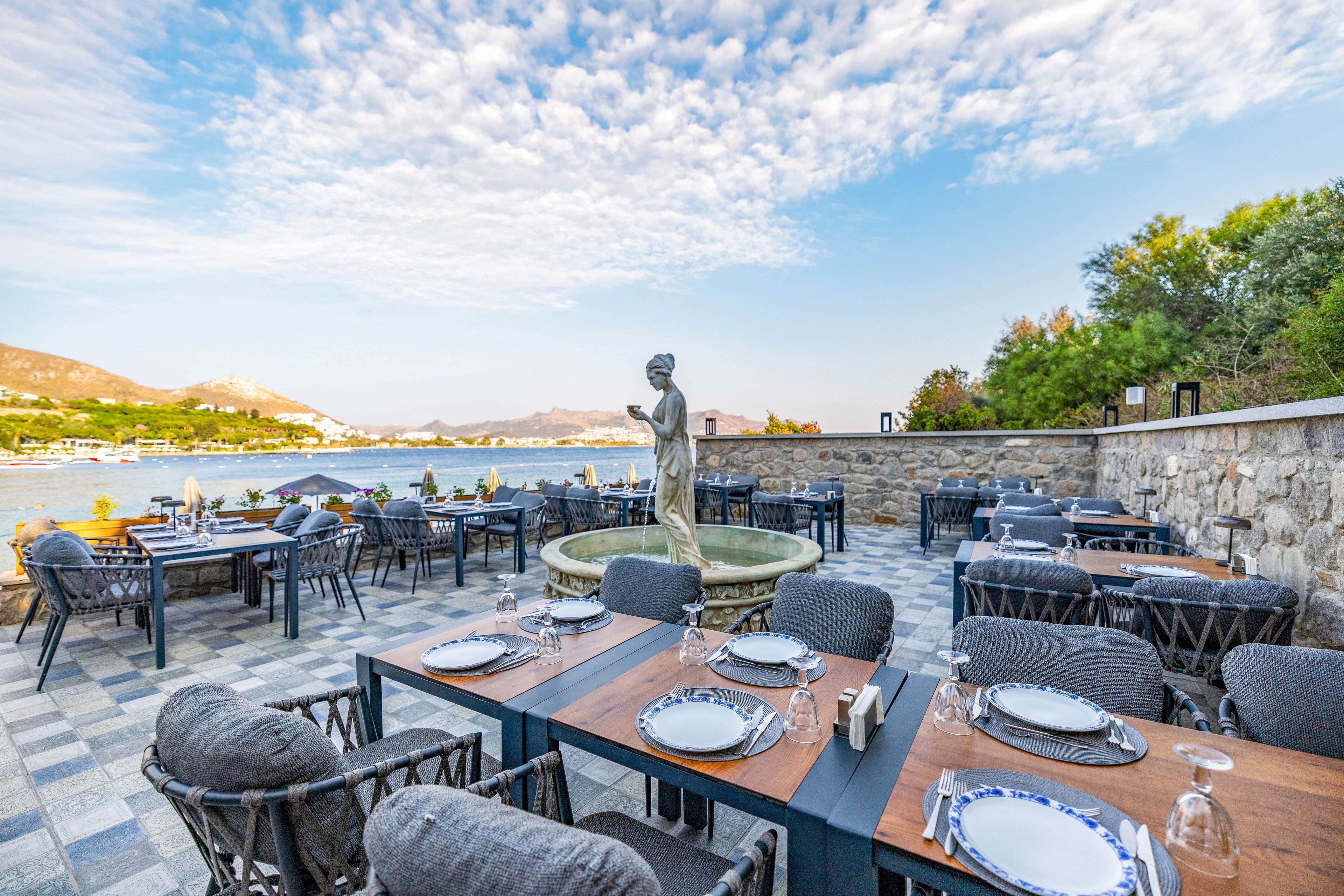 An elegant outdoor restaurant terrace features neatly set tables with plates, glasses, and cutlery, overlooking a scenic waterfront. A classical statue stands at the center of a stone fountain, surrounded by comfortable cushioned chairs. The setting is tranquil, with a stone wall, lush greenery, and mountains in the background under a partly cloudy sky.