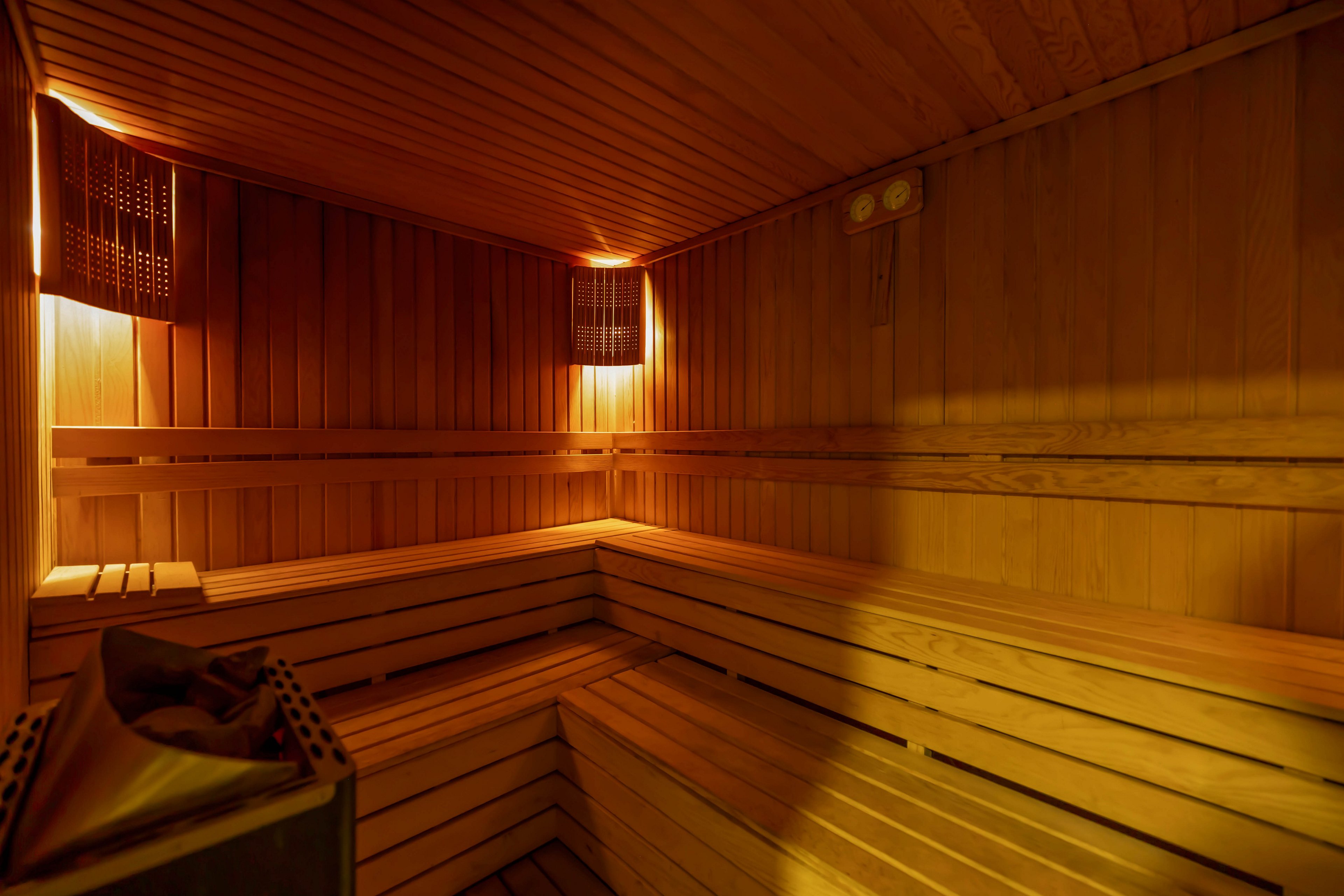 The image shows the interior of a traditional wooden sauna, featuring tiered bench seating and soft, ambient lighting. The space is empty, highlighting the clean lines of the wood paneling and the relaxing atmosphere. A heater is visible in the corner, and the overall mood is warm and inviting. No people or visible text are present in the scene.