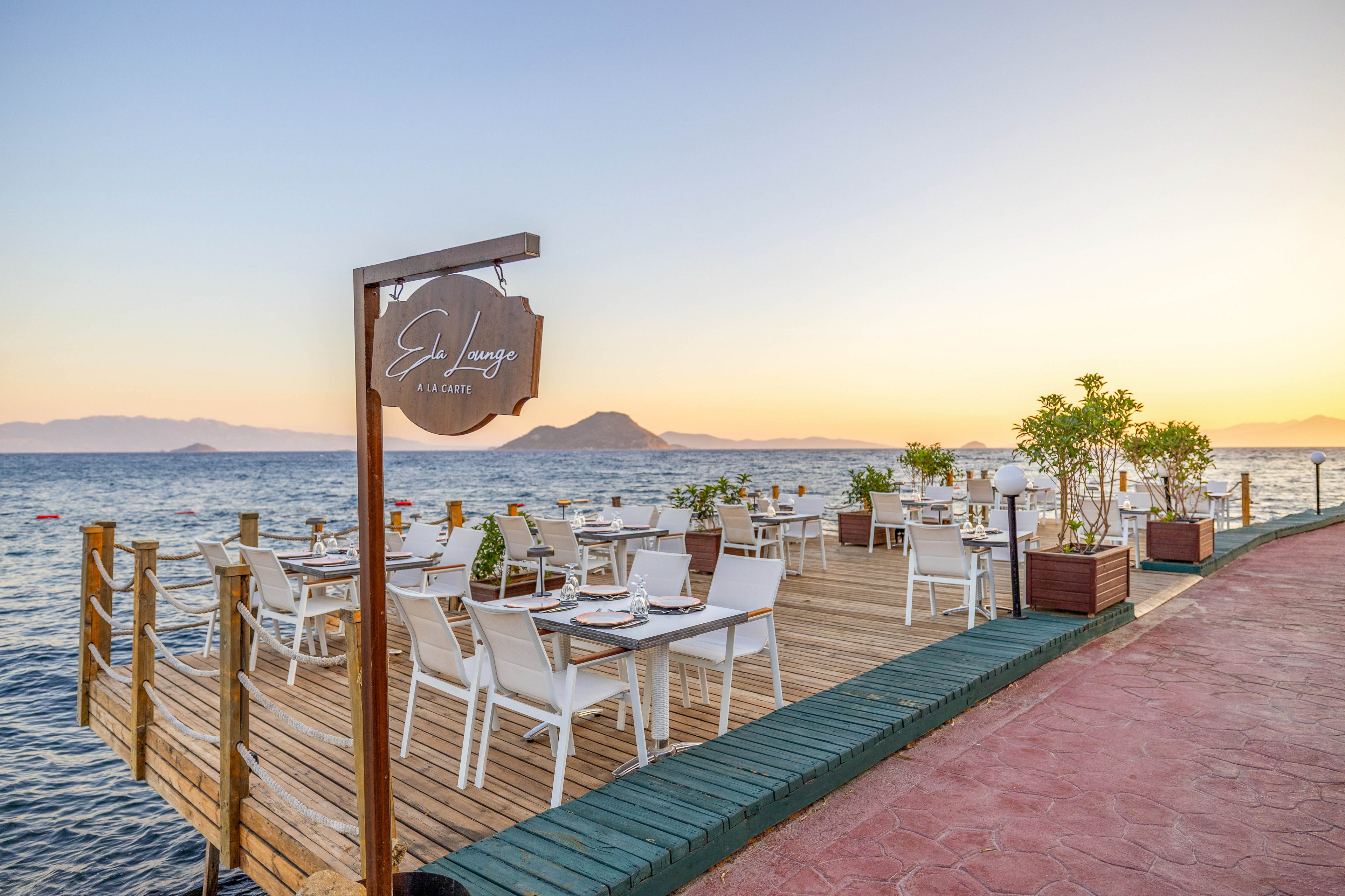 An outdoor restaurant terrace is set up on a wooden deck overlooking the sea at sunset. White tables and chairs are arranged neatly, with table settings ready for guests. The scene features calm water, distant mountains, potted plants, and a visible sign reading 'Ed Lounge Albatros.' The atmosphere is tranquil with warm, natural lighting.