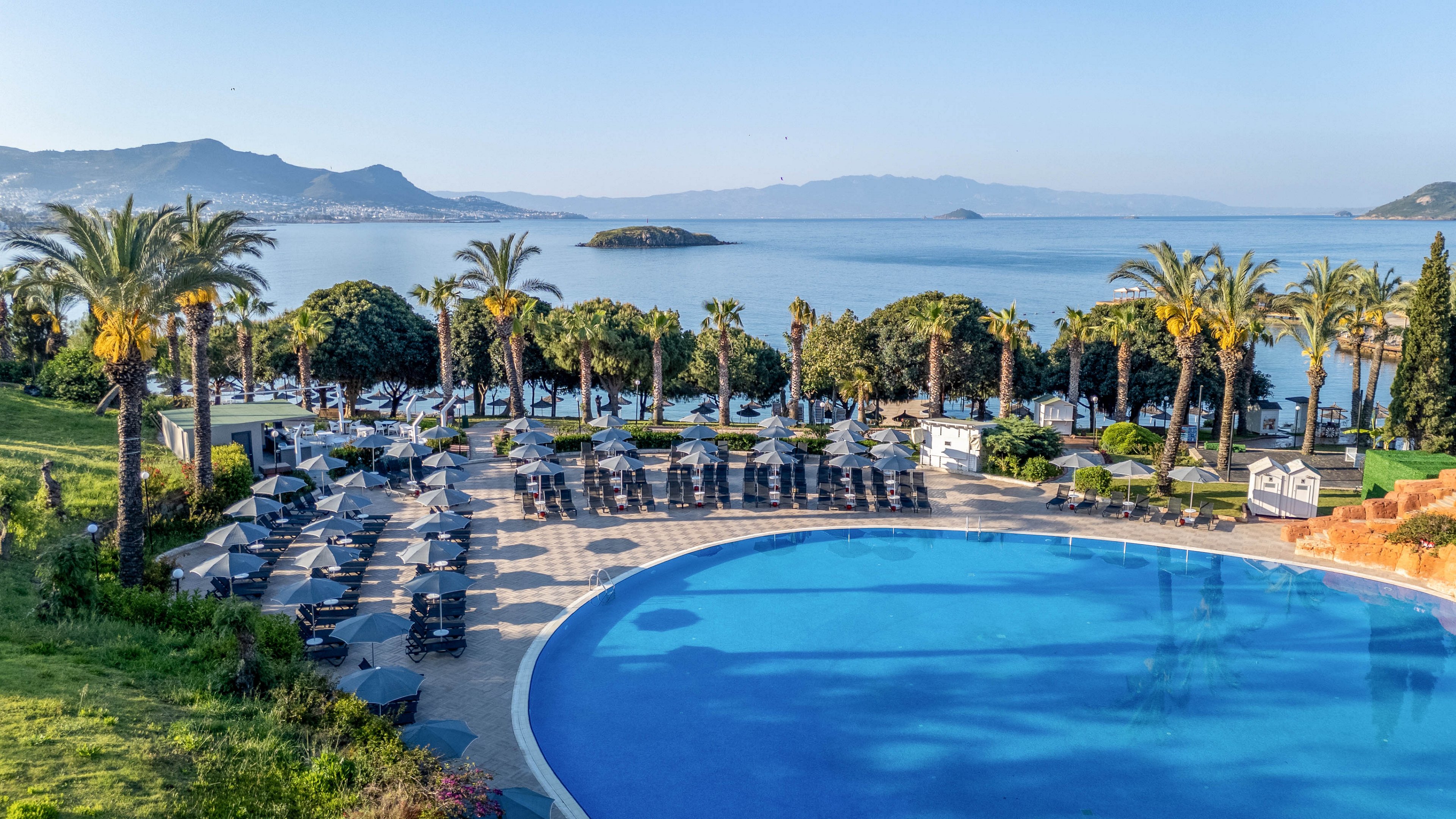 A large circular swimming pool is surrounded by numerous lounge chairs and umbrellas, set in a lush tropical landscape with palm trees. The scene overlooks a calm ocean with small islands and distant mountains, creating a serene vacation atmosphere. The setting is outdoors, under clear blue skies, with no visible people or text.