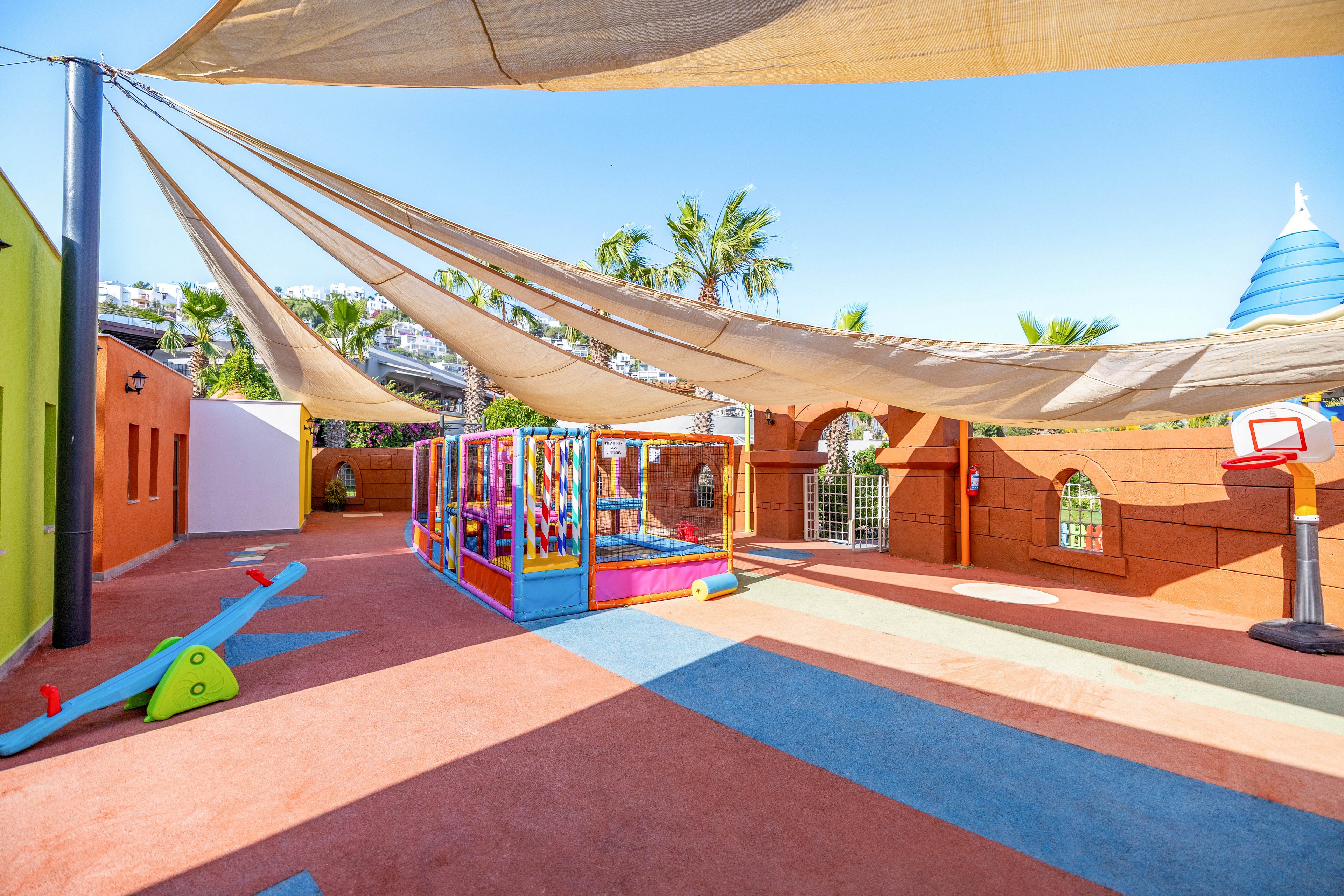 A vibrant outdoor playground features colorful play structures and soft flooring under large fabric sunshades. The area includes a small slide, climbing equipment, and a basketball hoop, all set against a backdrop of palm trees and bright walls. The scene is sunny and inviting, designed for young children to play safely. No visible text or numbers are present in the image.