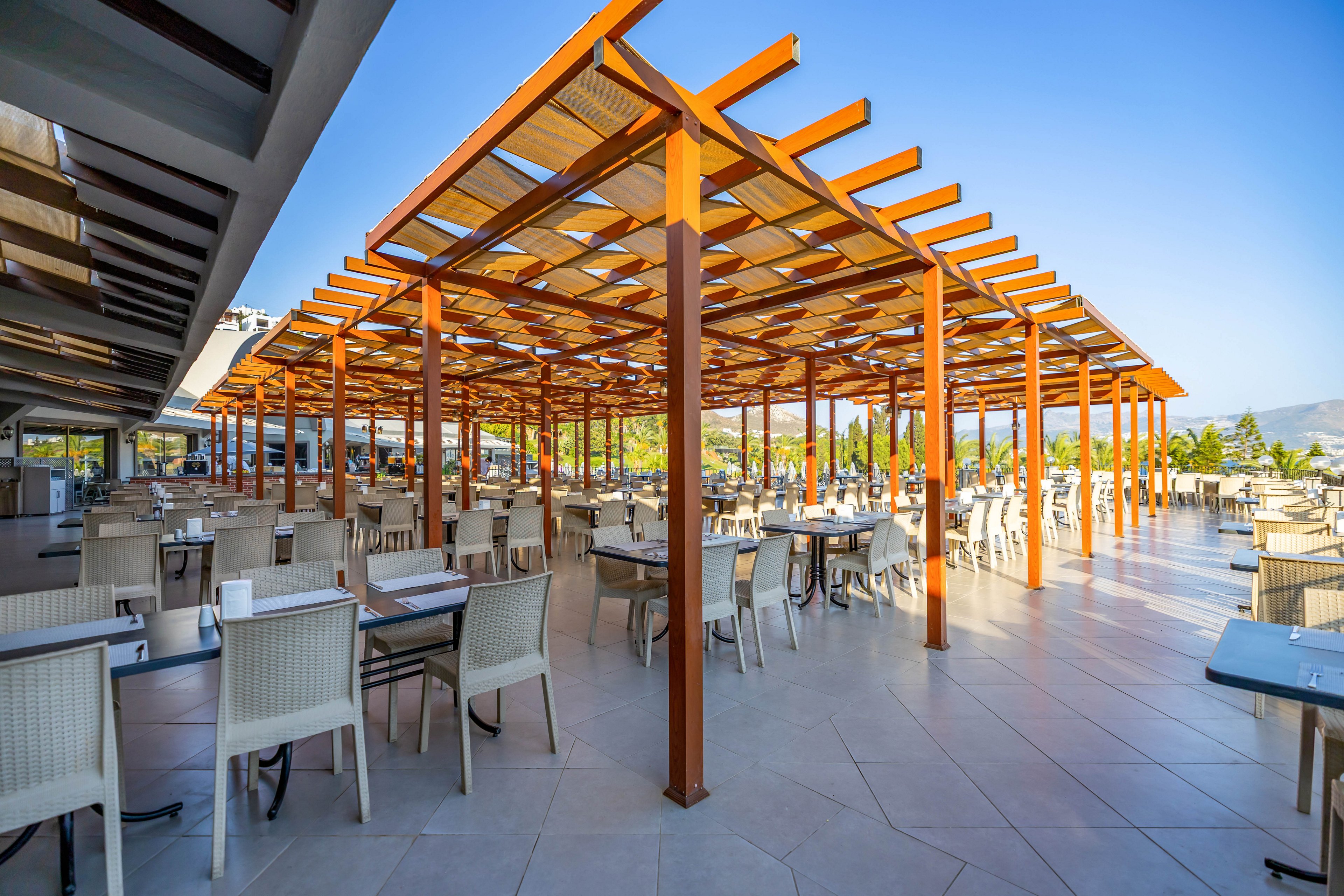 A spacious outdoor dining area features a modern wooden pergola structure providing partial shade over neatly arranged tables and chairs. The setting is bright and airy, with clear blue skies and distant greenery visible in the background. The scene is empty of people, emphasizing the clean, contemporary design and inviting atmosphere.