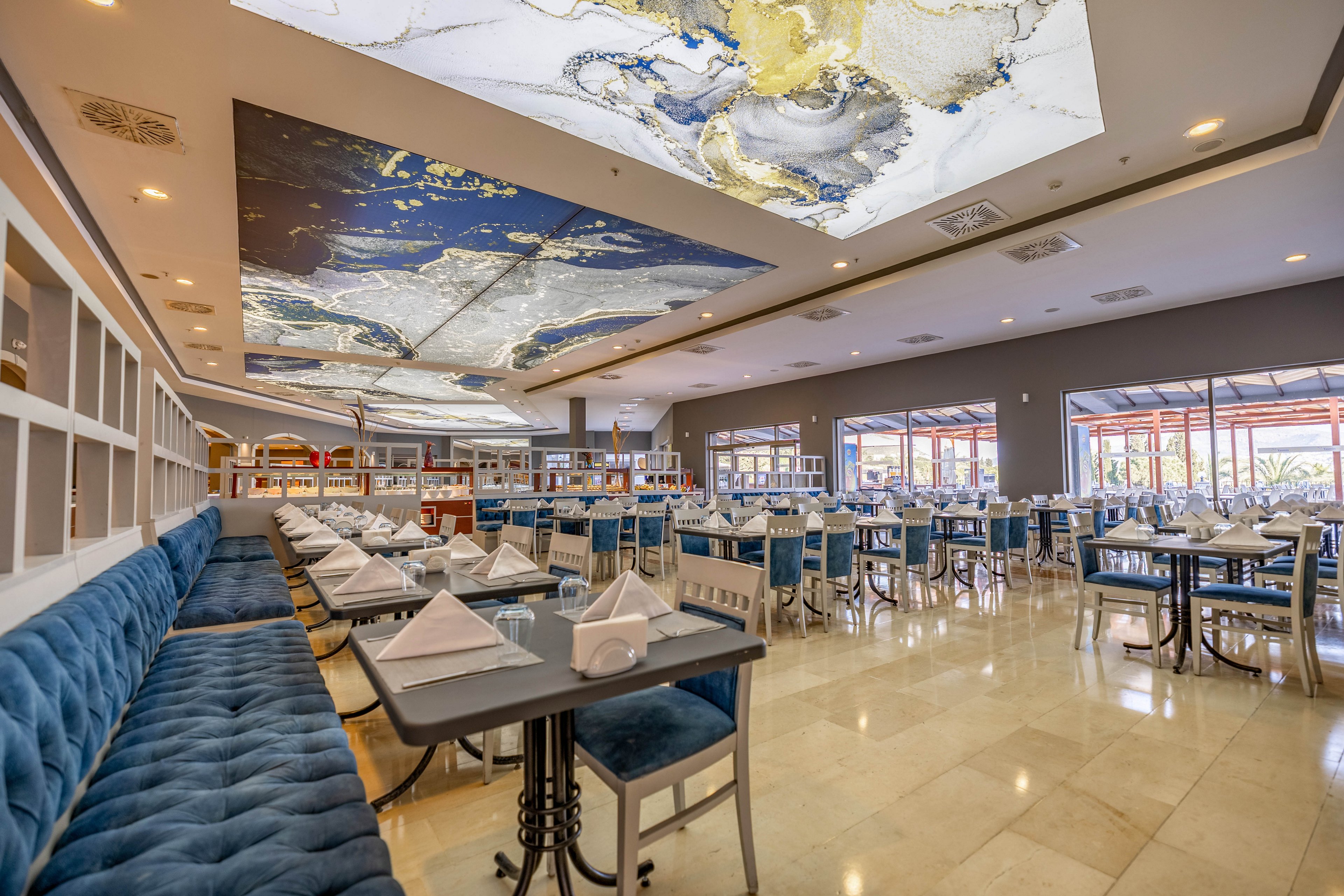 Spacious restaurant dining area featuring elegant blue velvet banquettes and neatly arranged tables with white napkins. The ceiling displays striking abstract artwork in blue, gold, and white tones, adding a contemporary touch. Large windows allow natural light to fill the room, highlighting the polished tile floors and inviting atmosphere. No visible people or text are present in the image.