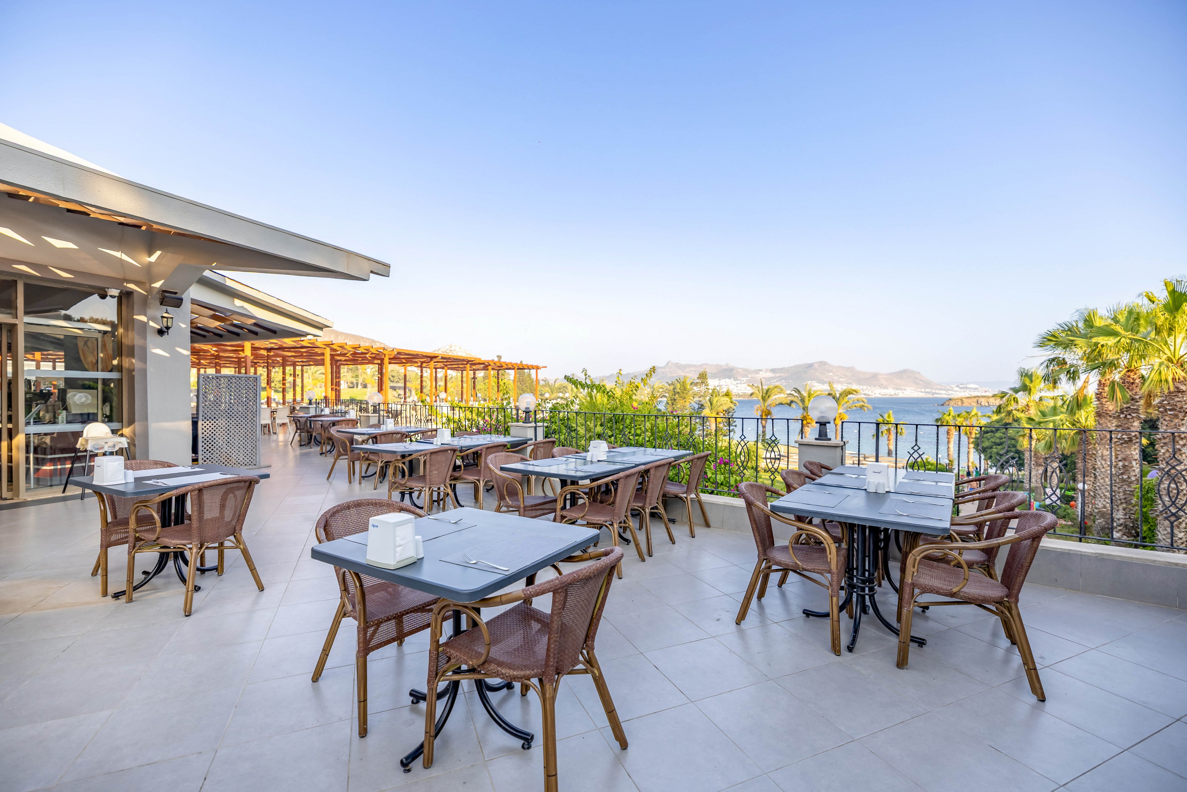 A spacious outdoor restaurant terrace features several empty tables and chairs arranged neatly under a clear blue sky. The setting overlooks a scenic coastal landscape with palm trees and distant mountains visible. The area is well-lit with natural sunlight, creating a relaxed and inviting atmosphere for dining. No people are present, emphasizing the tranquil and open environment.