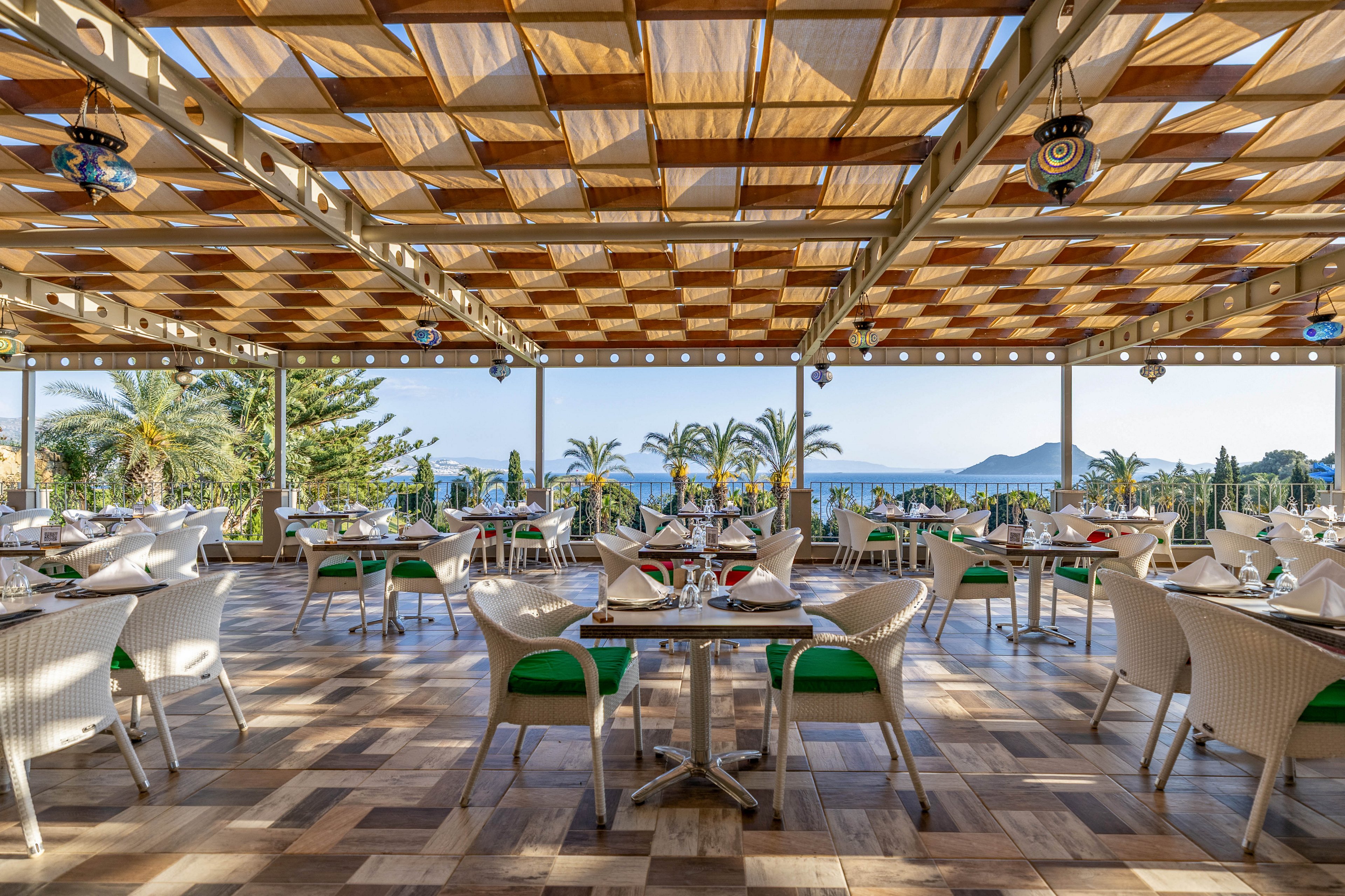 The image shows a spacious open-air restaurant with neatly arranged tables and chairs under a patterned wooden ceiling. The setting features a scenic view of palm trees and distant mountains, with the ocean visible in the background. The atmosphere is bright and inviting, with natural light and a relaxed coastal vibe. No visible text or numeric values are present in the image.