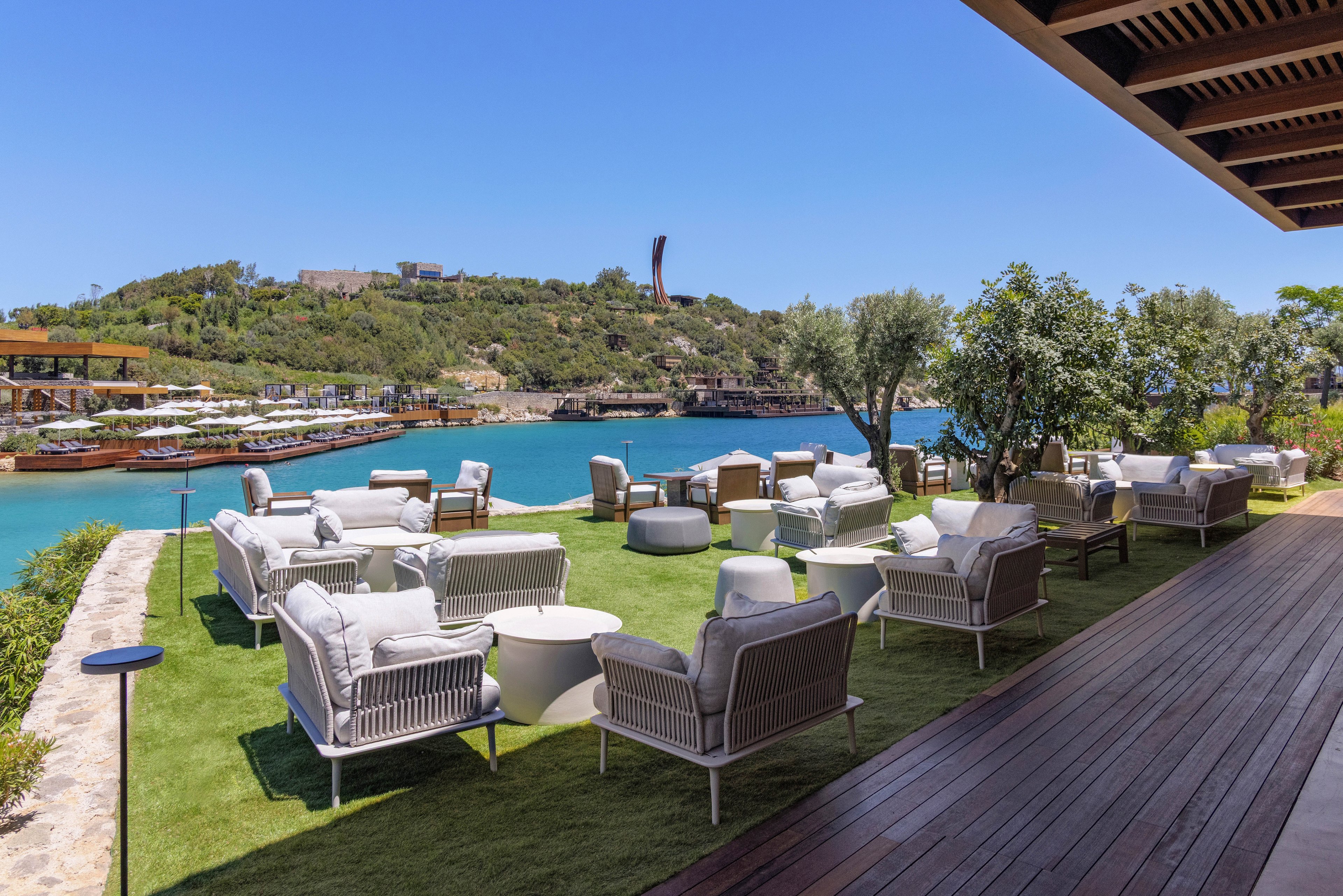 An upscale outdoor lounge terrace overlooks a calm turquoise bay. Modern white armchairs and sofas are arranged on a manicured grass area beside a wooden deck. In the background, sun loungers line the opposite shore and boats are docked along the water. Clear blue sky and lush green hills create a relaxed, resort-style atmosphere.