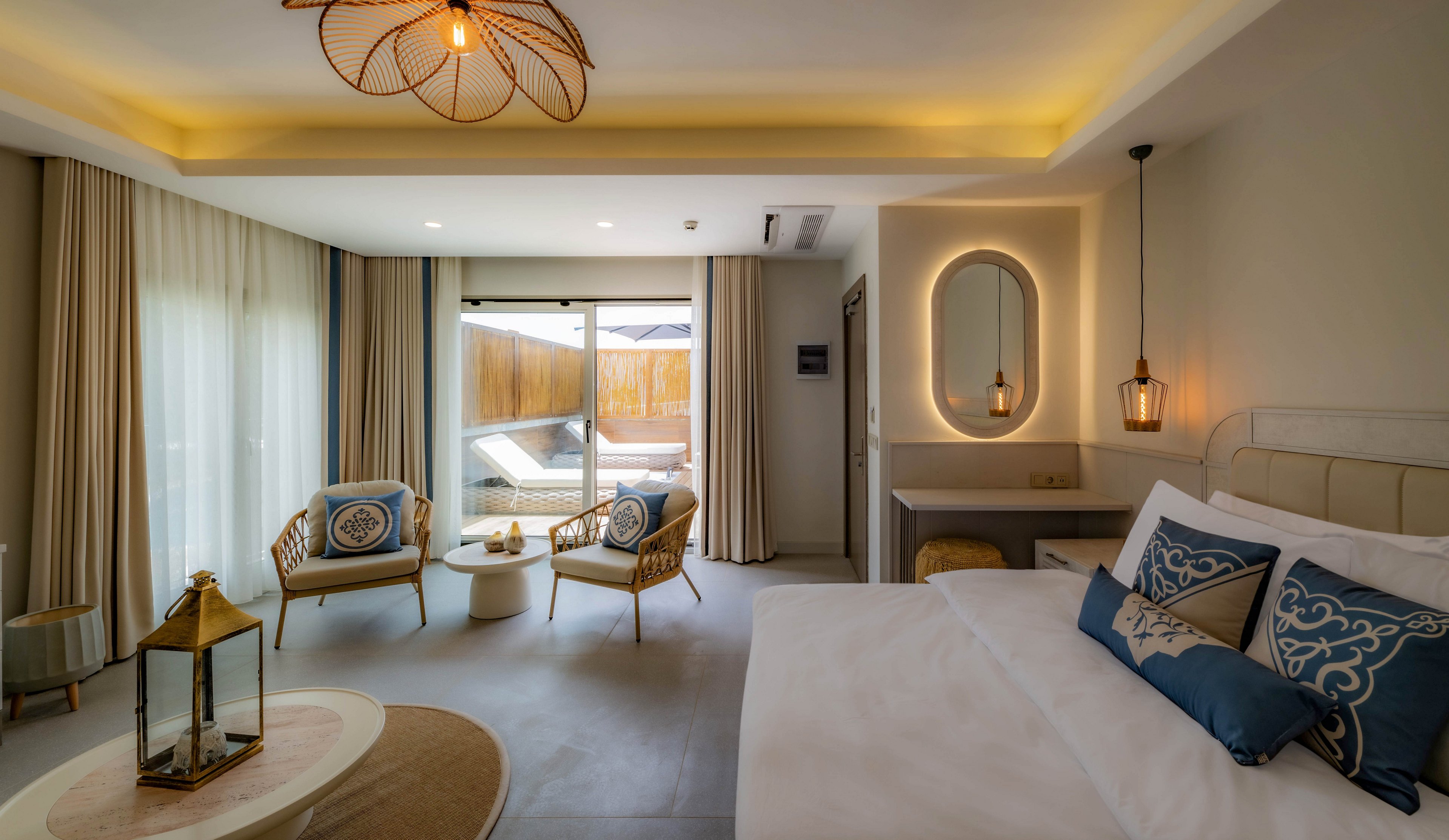 A spacious hotel suite features a large bed with decorative pillows, a stylish seating area with two chairs and a small table, and a lantern centerpiece. The room is illuminated by warm ambient lighting and natural light from floor-to-ceiling windows, which open to a private patio. Neutral tones and elegant accents create a relaxing and contemporary atmosphere.