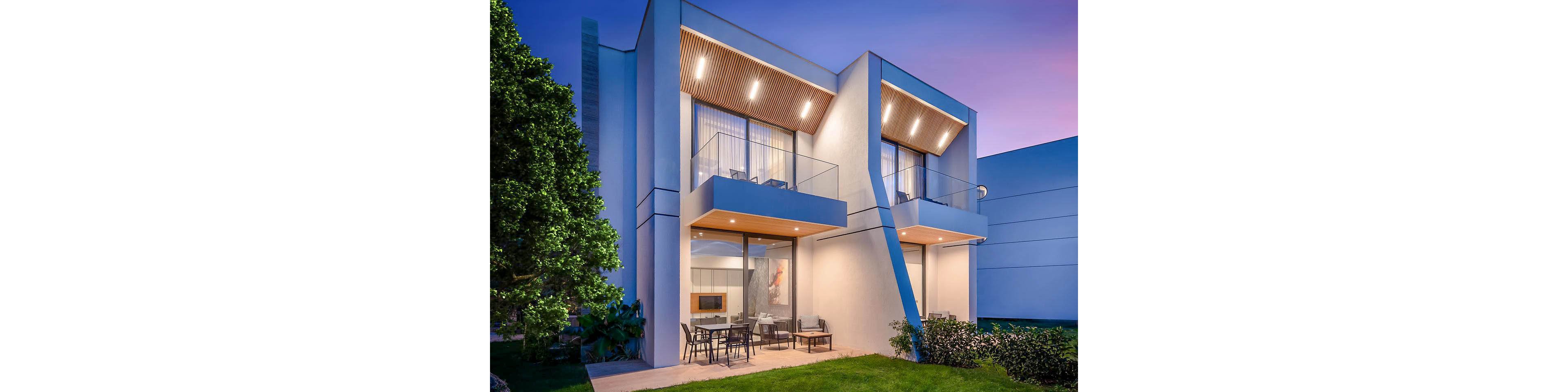 A contemporary two-story house with clean geometric lines is shown at dusk. Warm recessed lighting illuminates the balconies and covered patio area, contrasting with the cool blue evening sky. Large glass doors open onto an outdoor dining space on a neatly maintained lawn. Minimalist neighboring structures and a leafy tree frame the modern residential facade.