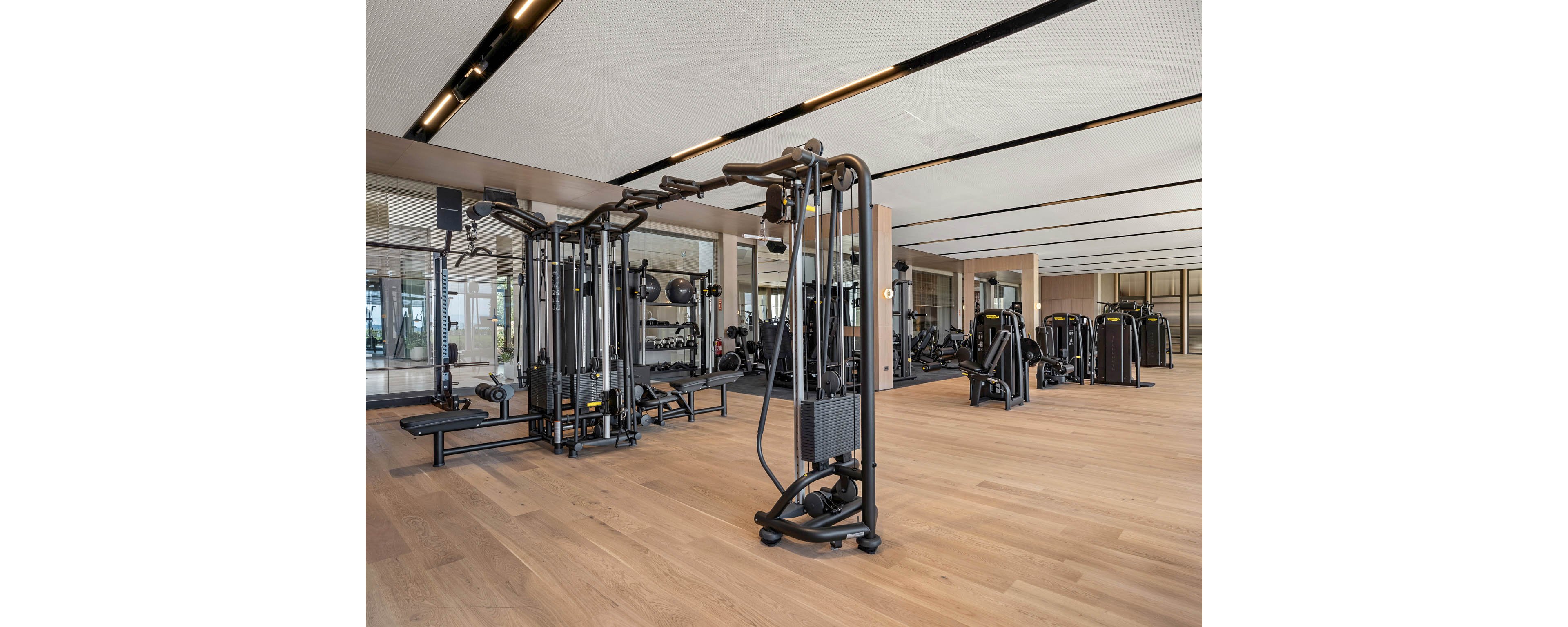 Spacious contemporary fitness center featuring multiple black strength training machines on light wood flooring. Large mirrors and glass walls reflect the equipment and create an open, airy atmosphere. The room is brightly lit with recessed ceiling lights and has a clean, minimalist interior design. No people are present, emphasizing the equipment and architectural details.