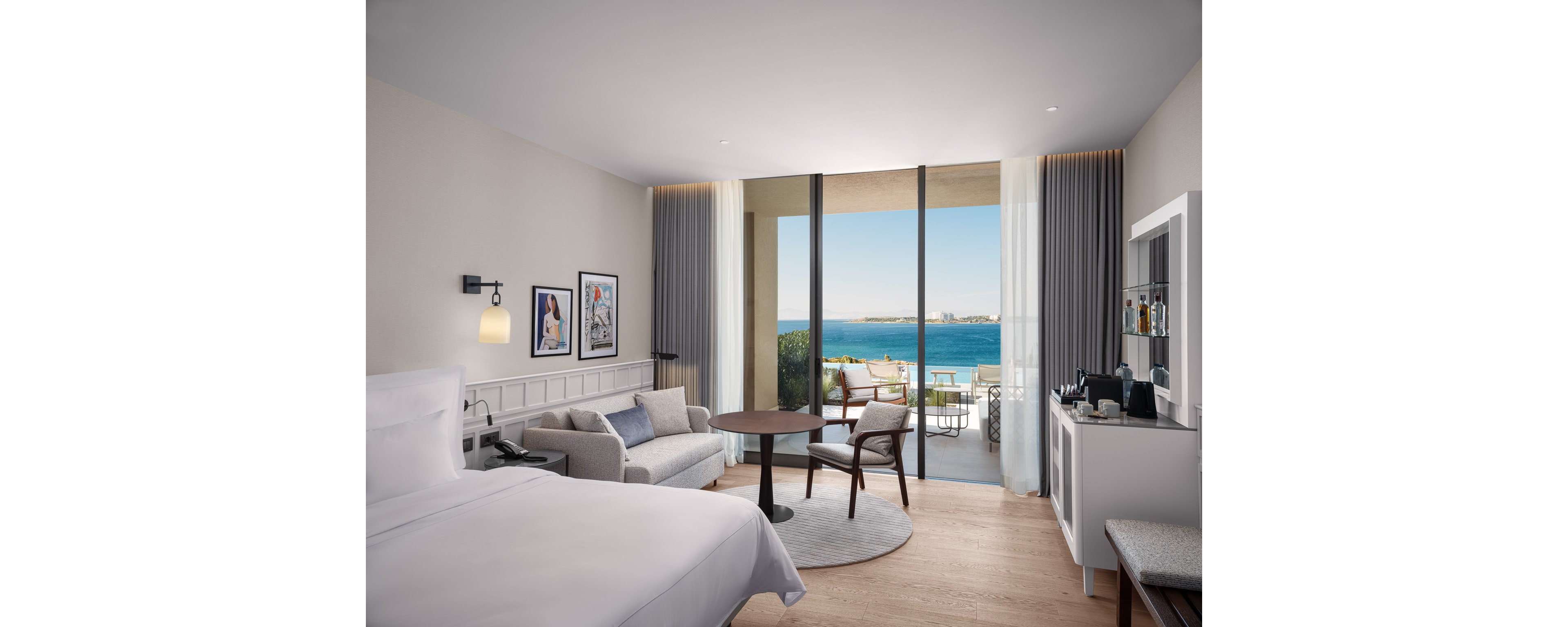 A contemporary hotel room features a neatly made bed and a cozy seating area with a sofa and round table. Floor-to-ceiling glass doors open onto a balcony overlooking a bright blue ocean and distant shoreline. Soft neutral tones, light wood flooring, and minimalist decor create a calm, upscale atmosphere. Wall art, sheer curtains, and a compact bar area add stylish details to the interior space.