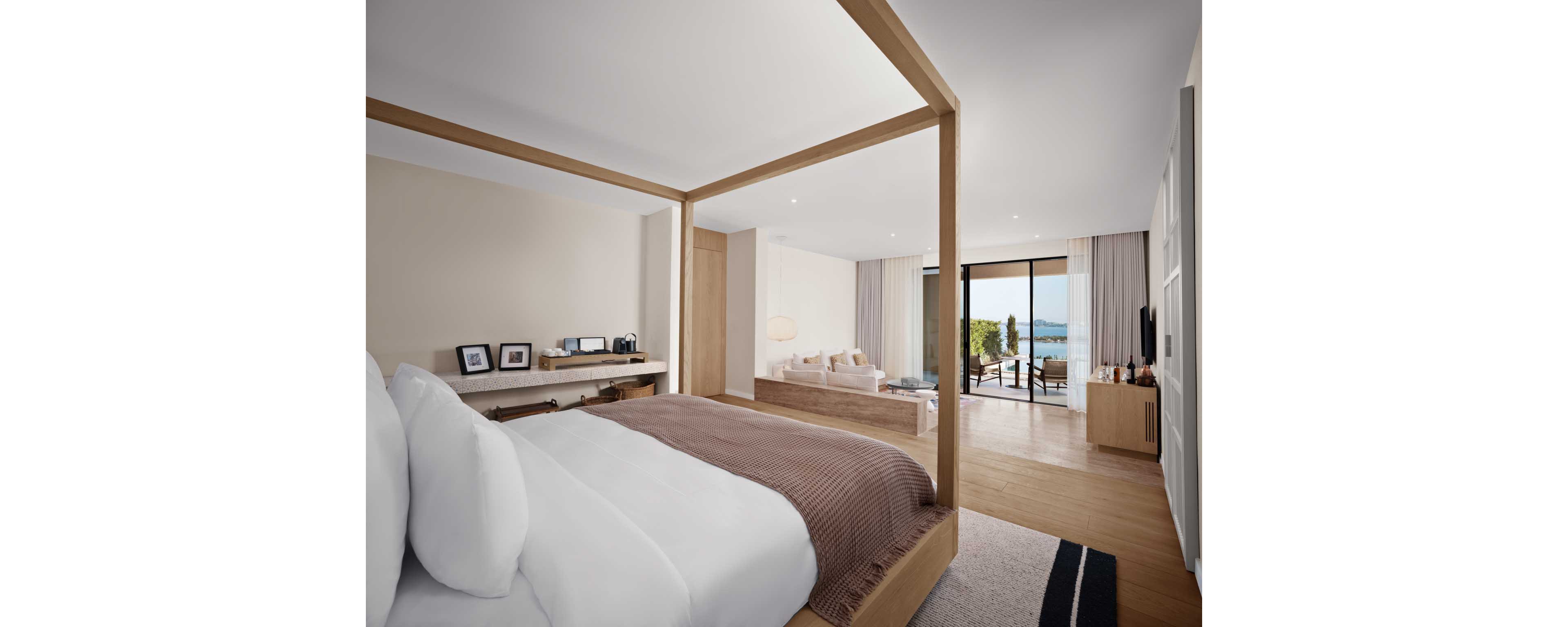 A contemporary hotel bedroom features a large bed with white linens and a brown throw, framed by a minimalist wooden canopy. The room extends into a bright sitting area with a sofa and coffee table, leading to a balcony through wide glass doors. Neutral tones, light wood flooring, and soft lighting create a calm, upscale atmosphere. Outside, the terrace overlooks water and greenery, suggesting a coastal or lakeside setting.