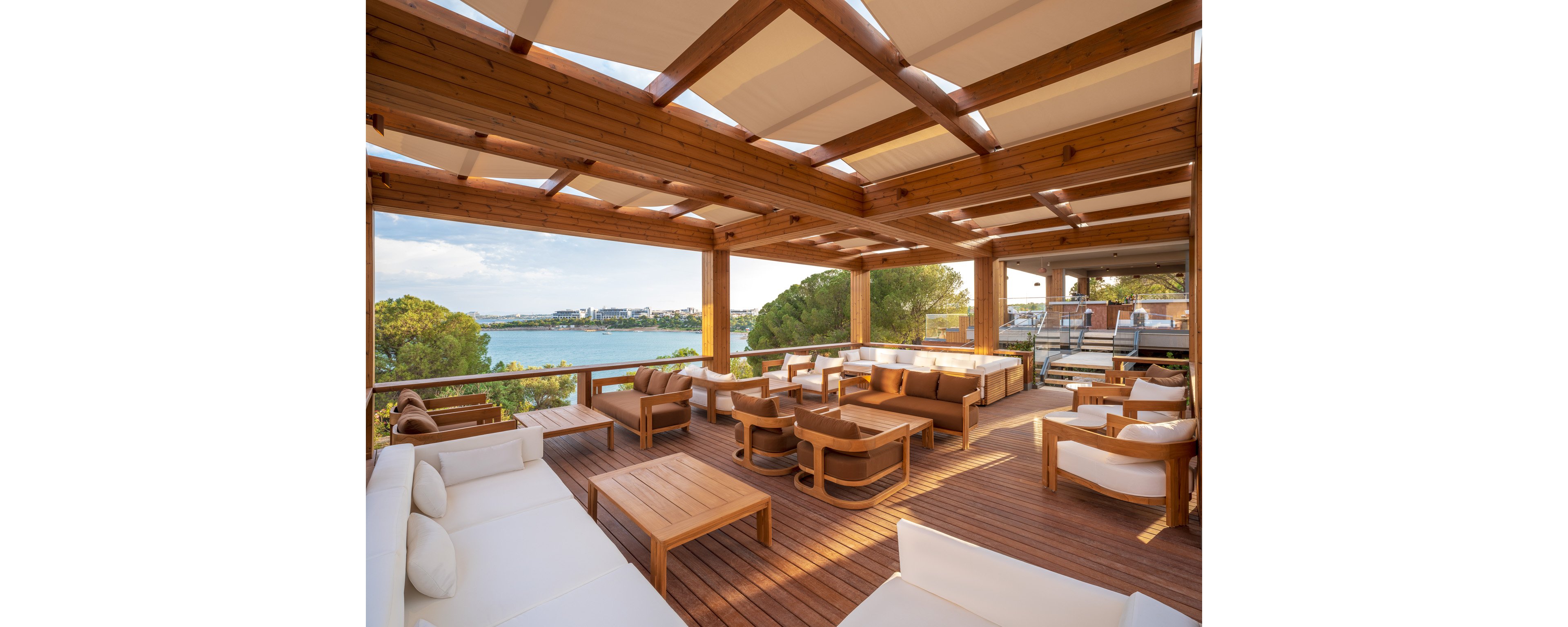 Spacious outdoor lounge area with wooden pergola and modern cushioned seating overlooks a calm waterfront. The terrace features multiple sofas, armchairs, and low tables arranged for relaxation and social gatherings. Warm wood tones contrast with white upholstery, creating a clean, contemporary coastal style. Trees and distant buildings frame the scenic view across the water under a clear sky.