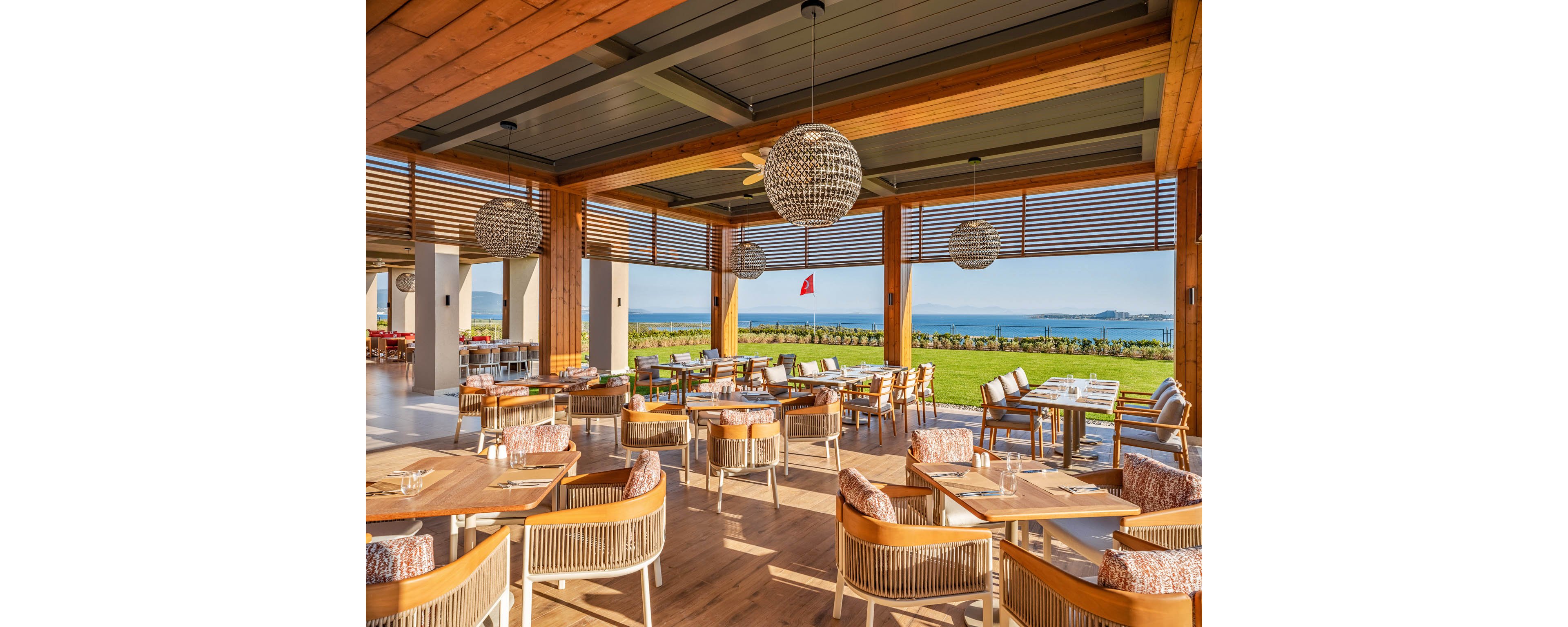 Spacious open-air restaurant with wooden tables and woven chairs overlooks a scenic coastline. Large spherical pendant lights hang from the ceiling, illuminating the neatly set dining area. Floor-to-ceiling openings with slatted shades frame a bright green lawn and blue sea beyond. A red flag is visible in the distance, adding a pop of color to the tranquil coastal setting.
