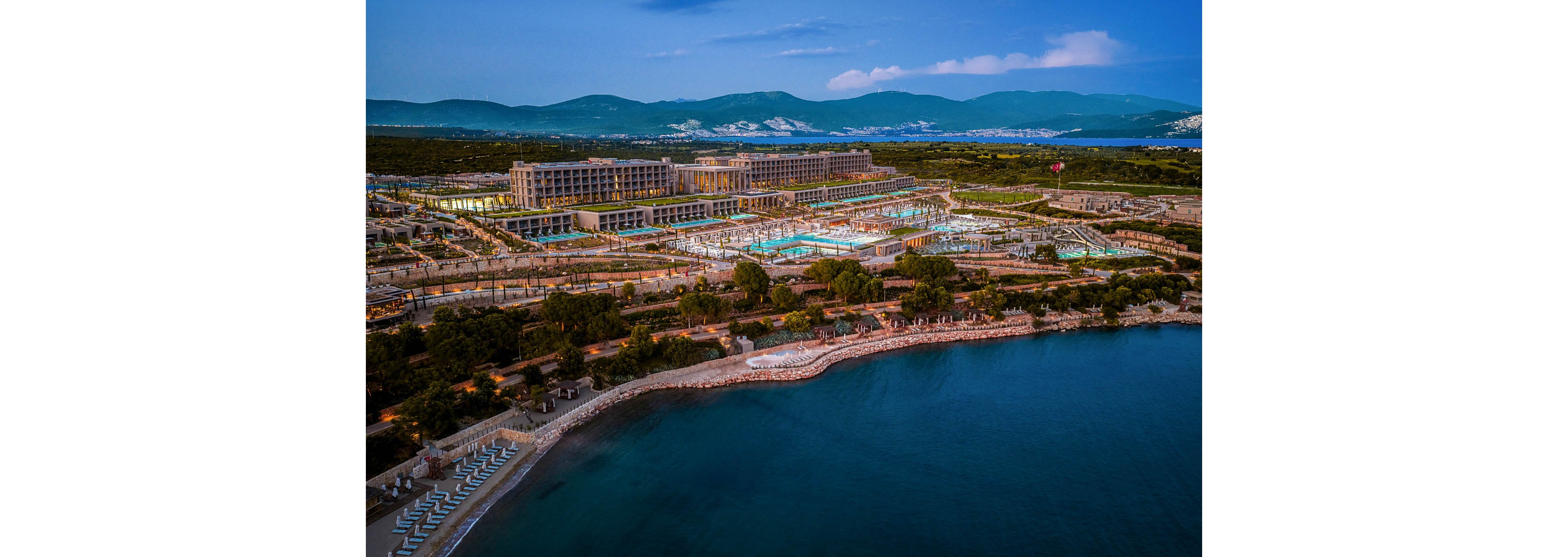 A large luxury resort complex is captured from above along a calm coastline at dusk. Multiple illuminated swimming pools and terraces stand out against the darker surrounding landscape. The property stretches parallel to the shoreline, with sun loungers arranged near the water and hills visible in the distance. The evening sky and soft lighting create a tranquil, inviting atmosphere.