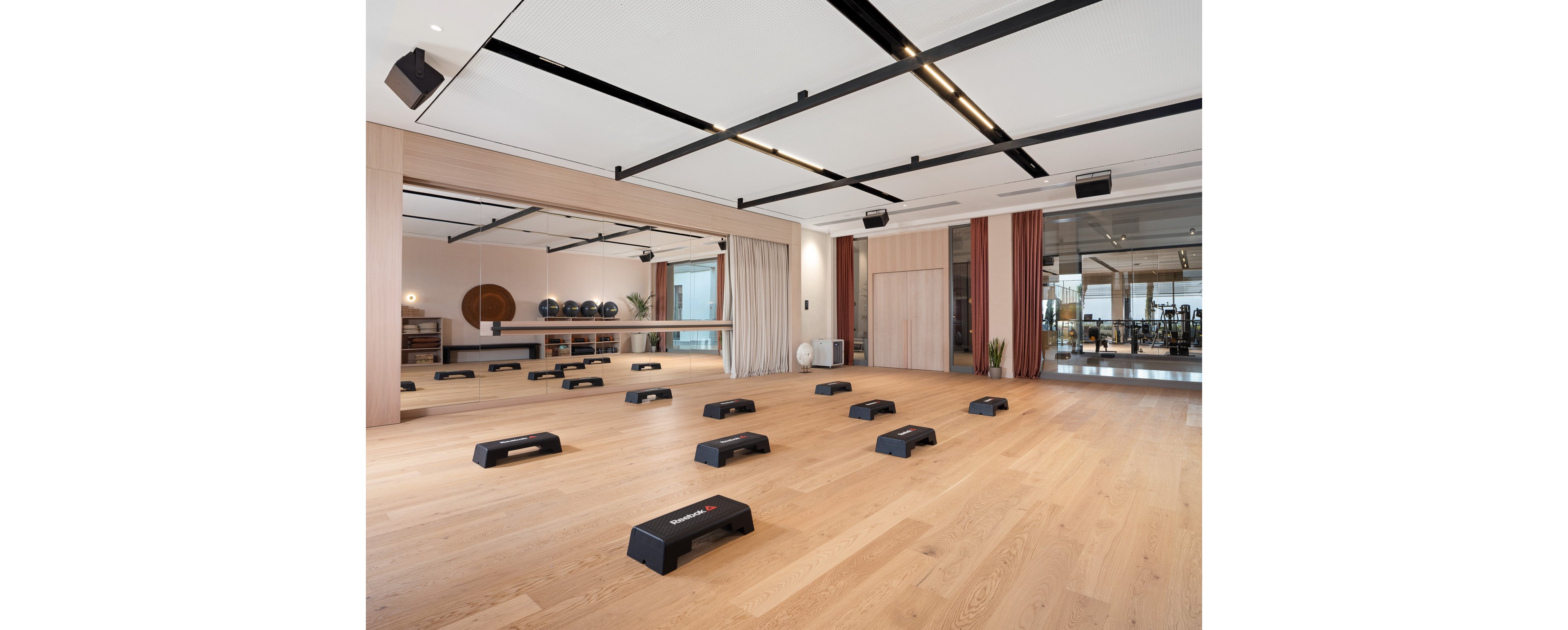 Spacious exercise studio featuring multiple black step platforms arranged on a light wooden floor. Large mirrors line one wall, reflecting the room and creating a sense of openness. In the background, shelves hold fitness balls and equipment, and glass doors reveal an adjacent gym area with machines. Neutral tones, clean lines, and soft lighting give the space a contemporary, minimalist feel.