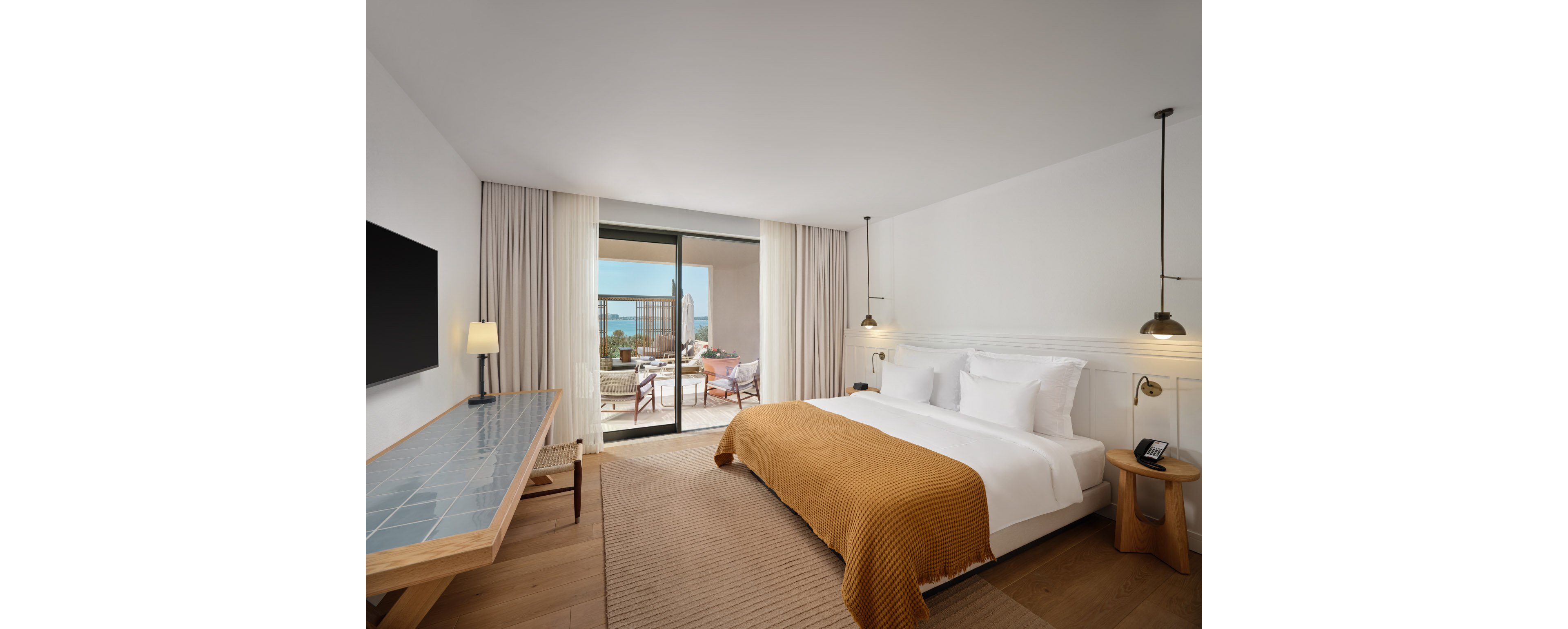 Contemporary hotel room features a large double bed with white linens and a mustard-colored blanket. A long wooden desk with a tiled surface and a wall-mounted flat-screen TV sit opposite the bed. Floor-to-ceiling curtains frame sliding glass doors that open to a furnished balcony overlooking the sea. Warm lighting and neutral tones create a calm, inviting atmosphere suitable for relaxation or business travel.