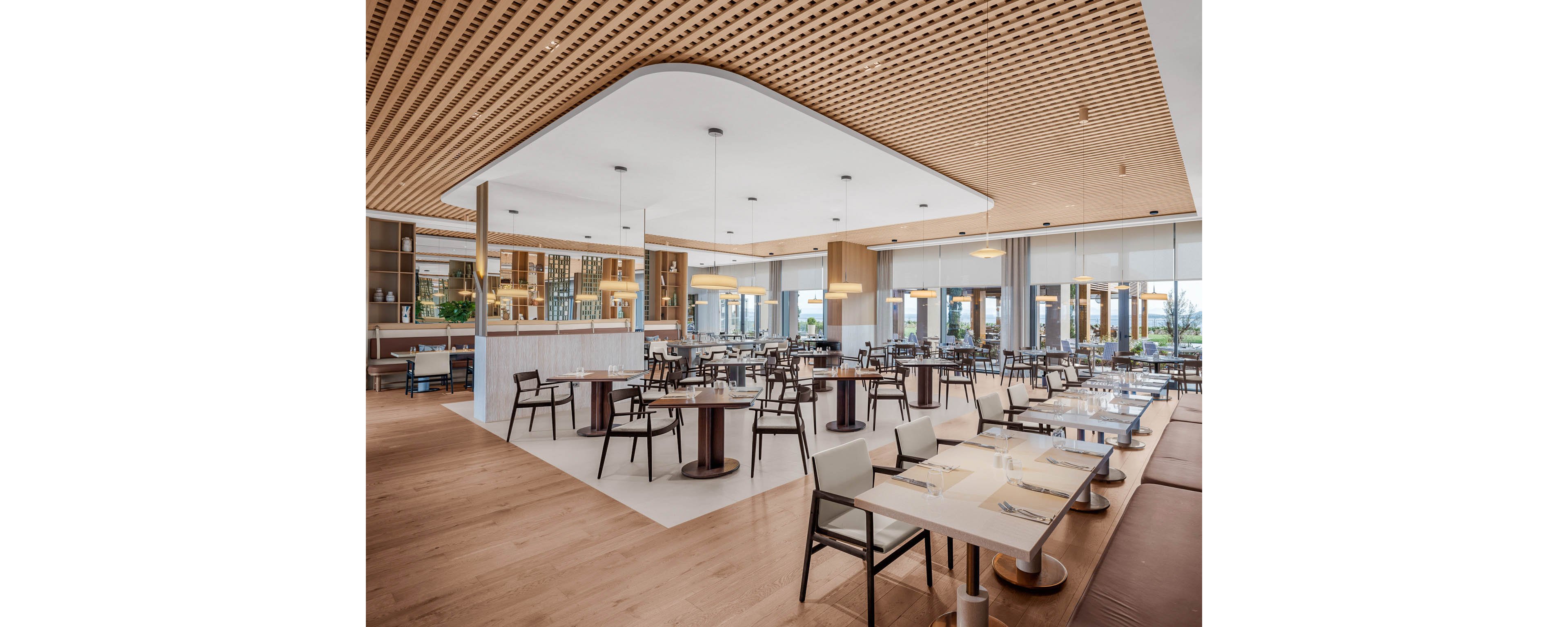 Contemporary restaurant dining area with neatly arranged tables and chairs is shown empty and ready for guests. The space features large floor-to-ceiling windows, allowing natural light to fill the room and highlight the clean, minimalist design. Wooden flooring and a patterned wood ceiling create a warm, inviting atmosphere complemented by soft pendant lighting. Table settings with plates, cutlery, and napkins emphasize a prepared, upscale dining experience.