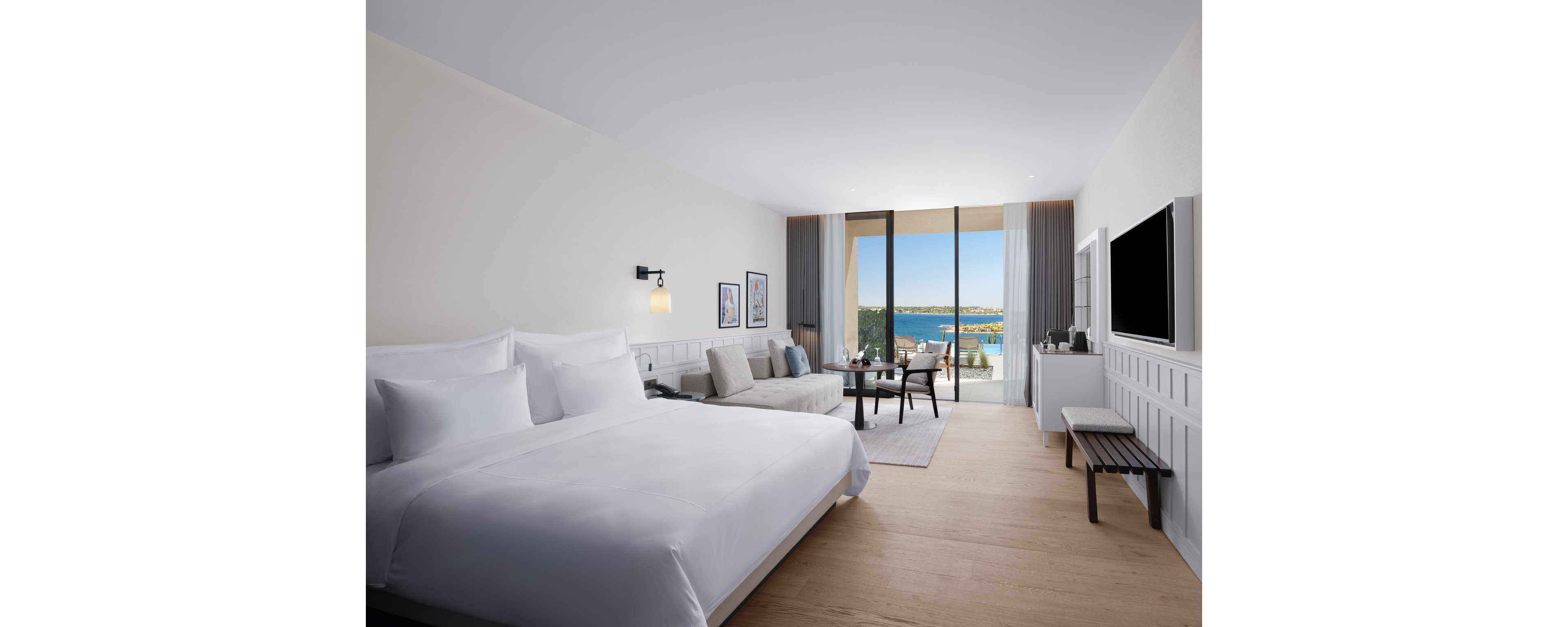 A spacious modern hotel room features a large neatly made bed with white linens and a light wood floor. A sofa seating area and round table sit near floor-to-ceiling glass doors that open to a balcony overlooking the sea. Minimalist decor includes framed artwork, a wall-mounted television, and subtle built-in cabinetry. Natural light fills the room, creating a bright and relaxing coastal atmosphere.