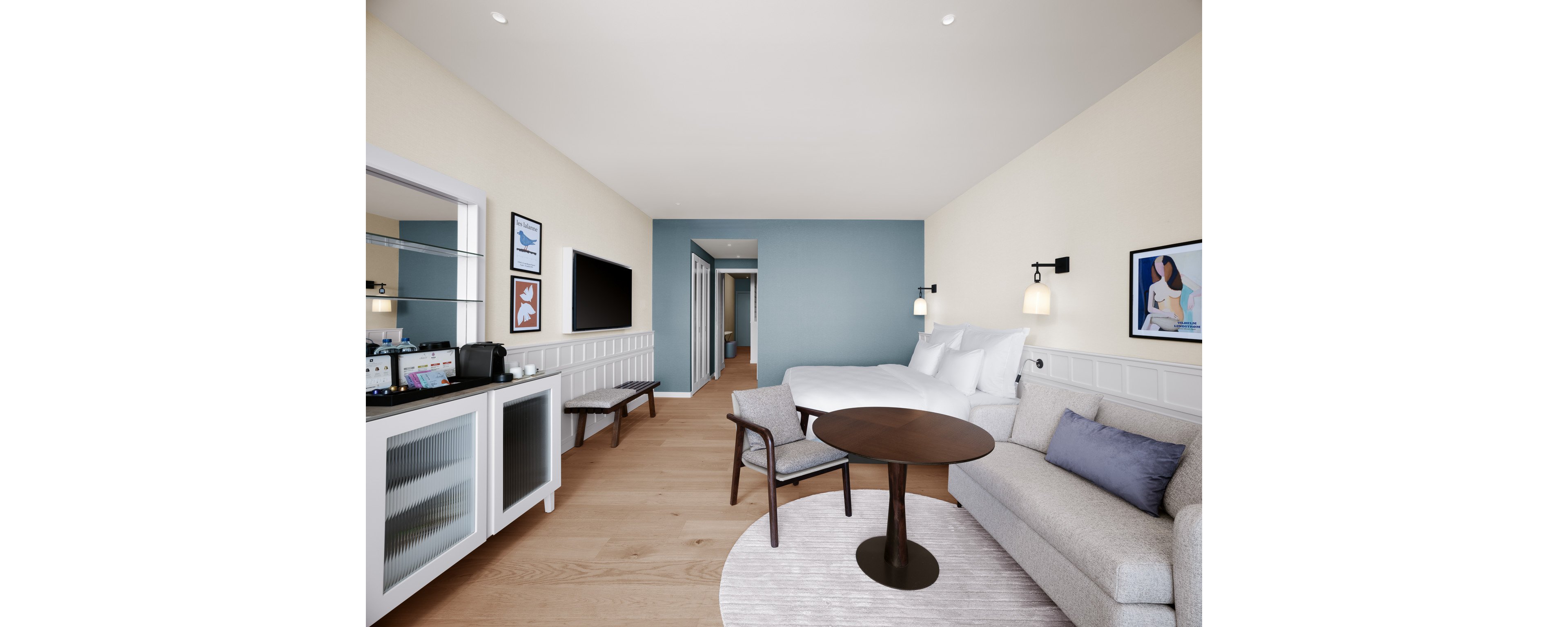 Bright contemporary hotel room featuring a large bed against a blue accent wall. A cozy seating area with a round table, armchair, and light-colored sofa sits on a circular rug. Built-in cabinetry with a minibar, coffee station, and flat-screen TV lines one wall. Soft neutral tones, wood flooring, and framed artwork create a calm, inviting atmosphere.