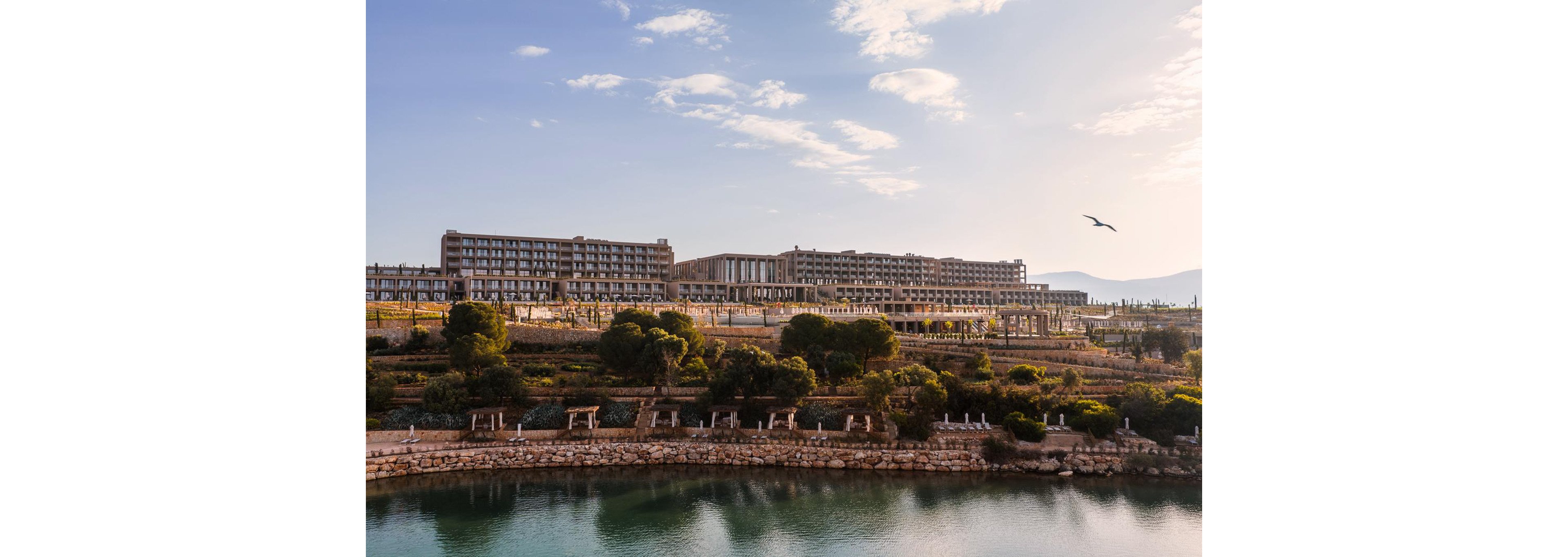 A large resort or hotel complex stretches across the horizon, overlooking a calm body of water. Terraced stone walls and lush green trees line the waterfront, creating a layered landscape. The sky is clear with soft clouds, and a single bird is captured in flight above the buildings. Distant mountains are faintly visible, adding depth and a sense of tranquility to the scene.