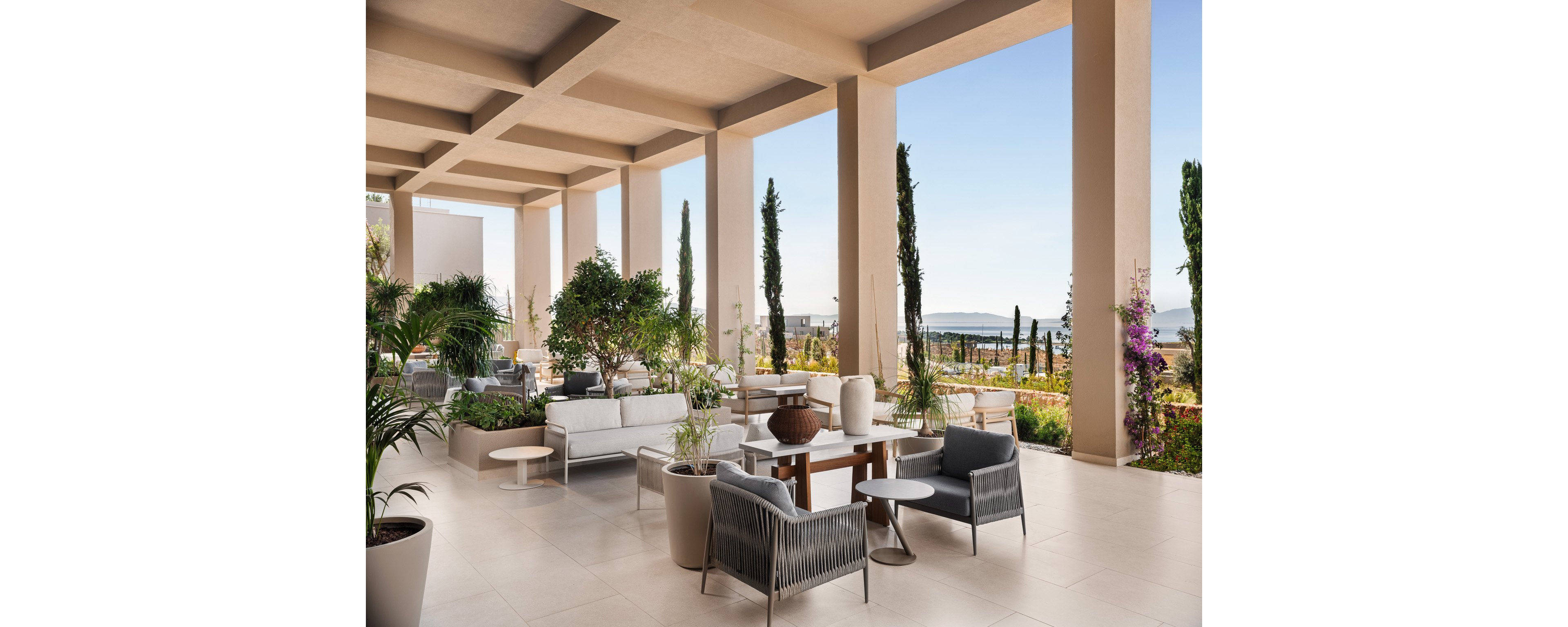 A contemporary open-air terrace features stylish lounge seating and low tables arranged beneath a geometric ceiling. Potted trees and lush greenery soften the clean architectural lines and frame views of the distant coastline and sea. Neutral tones of beige and gray dominate the space, complemented by natural textures and minimalist decor. The scene evokes a relaxed, upscale atmosphere ideal for outdoor socializing or quiet relaxation.
