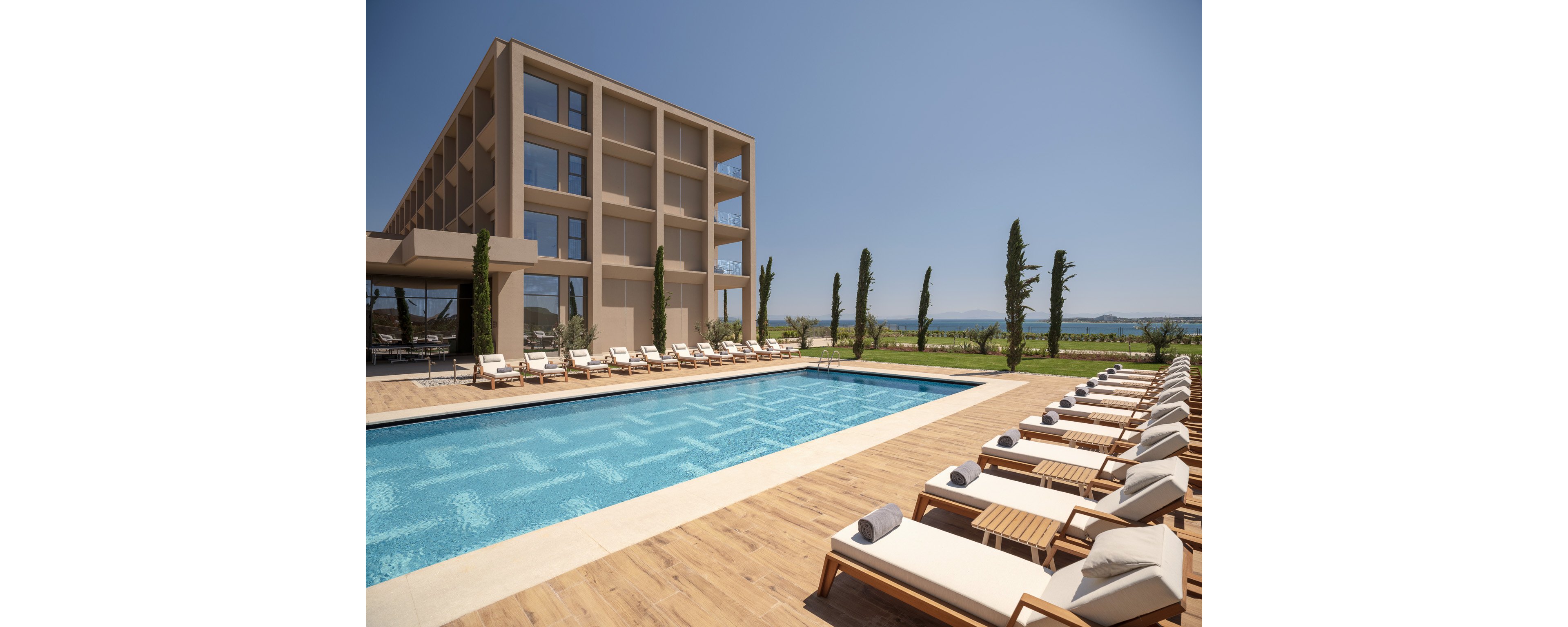 A contemporary multi-story hotel overlooks a large rectangular outdoor swimming pool. Numerous sun loungers with white cushions and rolled towels line the wooden deck on both sides of the pool. Tall, slender trees and manicured lawns frame the leisure area, with distant hills and water visible under a clear blue sky. The scene suggests a calm, sunny vacation atmosphere at a modern resort.
