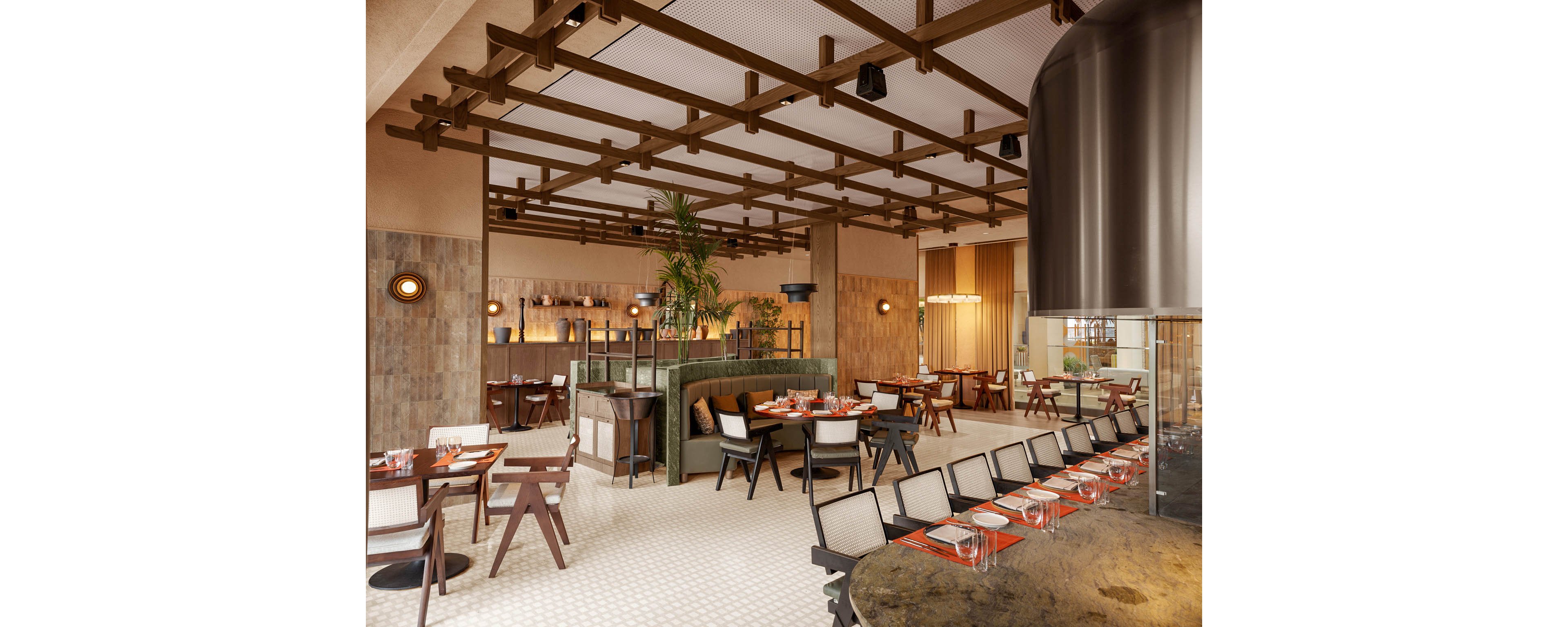 A contemporary restaurant dining area features neatly arranged tables and chairs with red place settings. The space includes a central seating section with green upholstered benches and decorative plants. Warm wooden beams on the ceiling, textured walls, and soft lighting create a cozy, inviting atmosphere. The overall design blends modern furniture with natural materials for a stylish, upscale look.