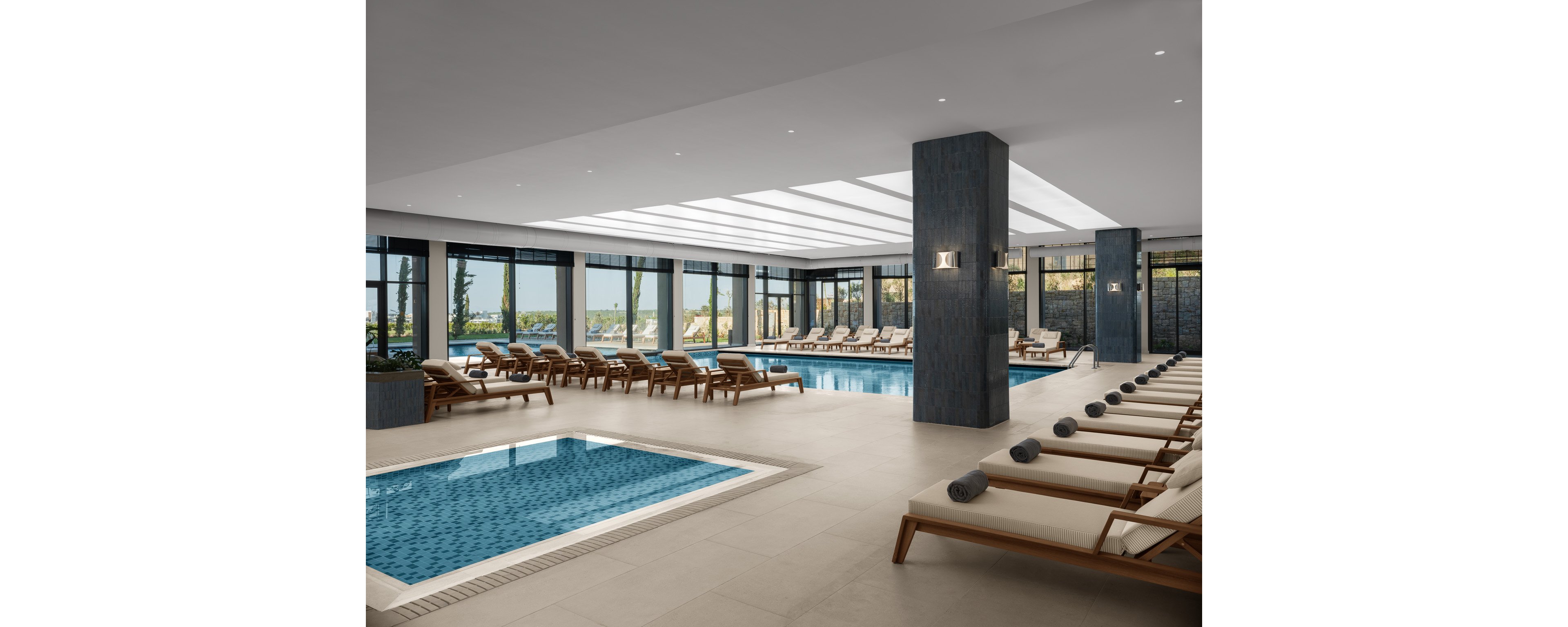 Spacious indoor pool area features a large rectangular swimming pool and a smaller adjacent spa pool. Numerous cushioned sunbeds with rolled gray towels line both sides of the room, creating a resort-style relaxation space. Floor-to-ceiling windows reveal an outdoor landscape and allow natural light to complement the bright skylight ceiling. Clean lines, neutral tones, and blue water accents give the scene a calm, contemporary spa atmosphere.
