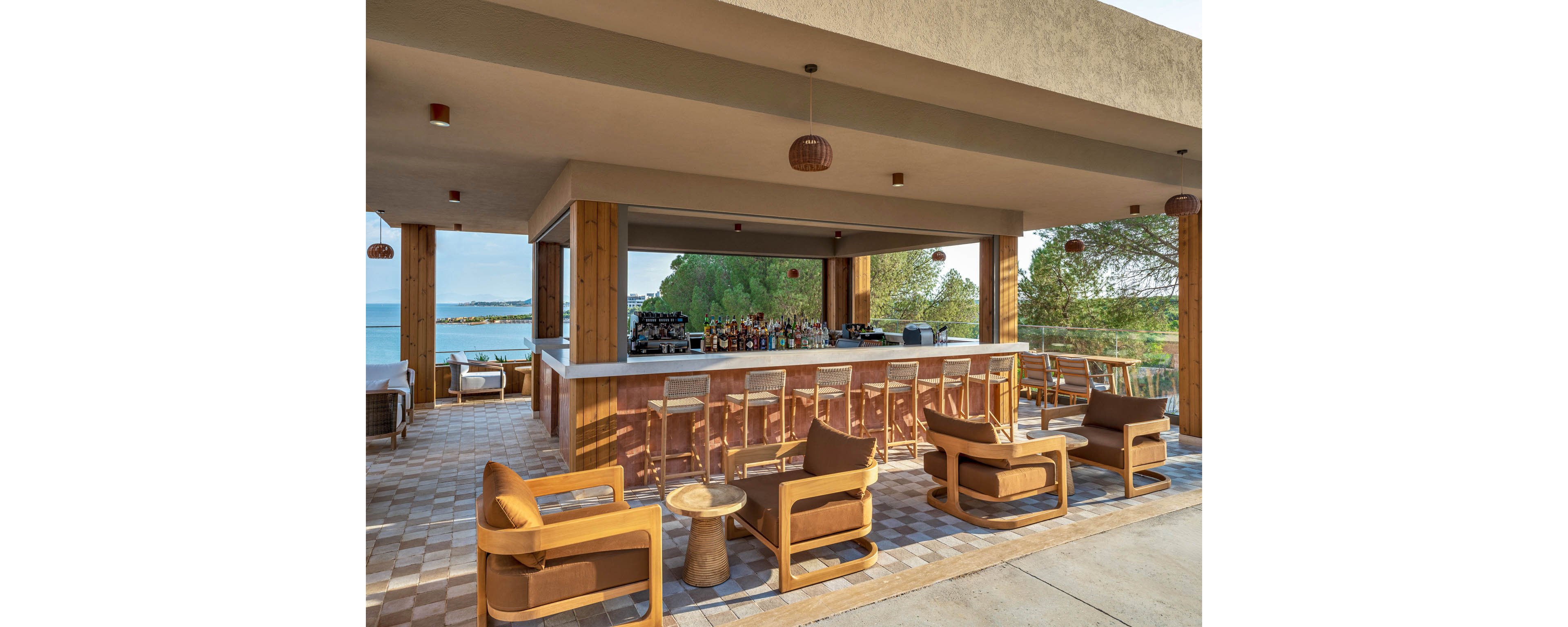 An open-air bar with wooden counters and cushioned lounge chairs sits under a modern canopy. The setting overlooks a scenic coastline with calm blue water and lush green trees in the background. Bottles and bar equipment line the counter, while barstools face the ocean view. Warm earth tones and natural materials create a relaxed, contemporary resort atmosphere.