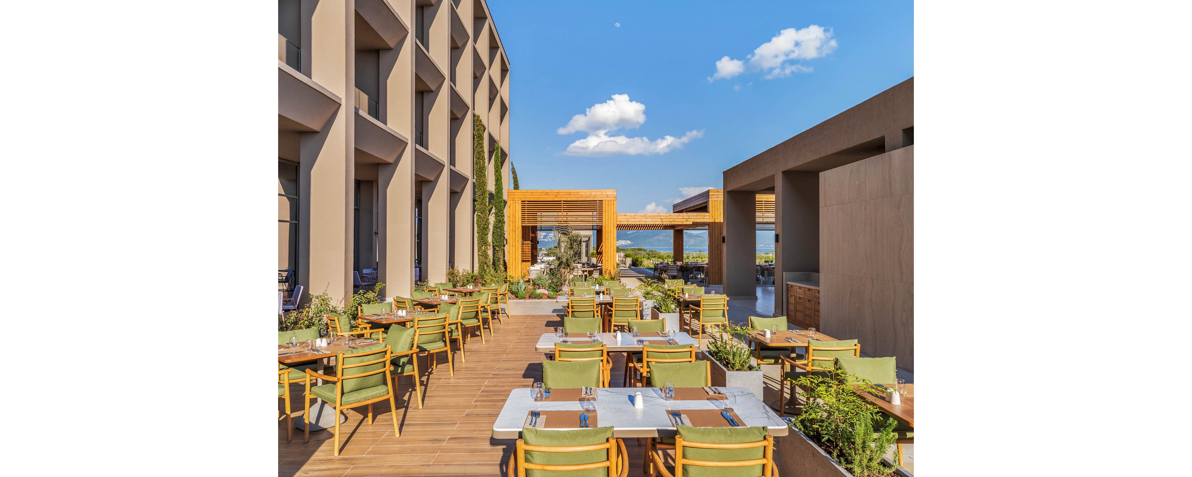 A contemporary outdoor restaurant terrace features neatly arranged tables and green cushioned chairs on a wooden deck. The setting is framed by modern architectural buildings and pergola-style wooden structures, creating a stylish open-air dining area. Small plants and greenery line the edges of the terrace, adding a natural touch to the clean design. A bright blue sky with scattered clouds enhances the inviting, relaxed atmosphere of the space.