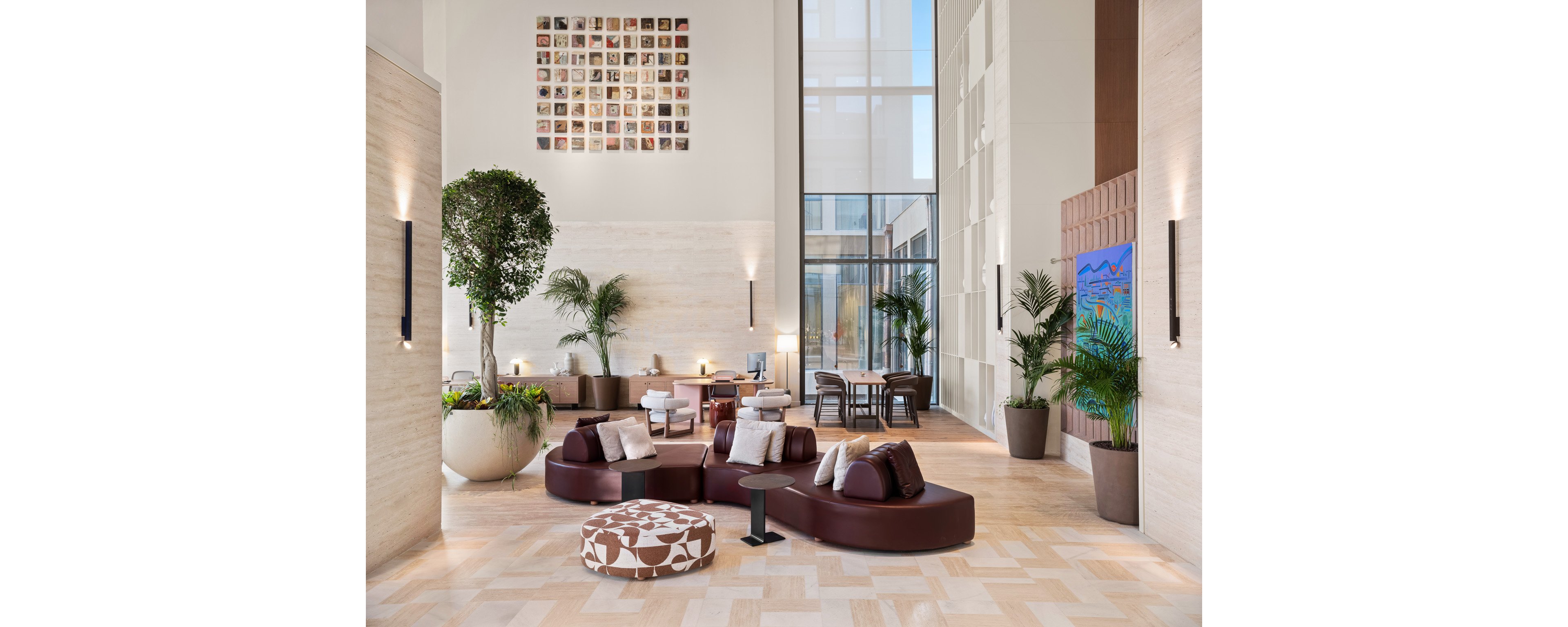 A spacious, modern hotel lobby features curved brown leather seating with assorted cushions and a patterned ottoman. Tall potted plants and trees add greenery along the light stone walls and floor. Large windows at the back bring in natural light and reveal an outdoor area with tables and chairs. Wall-mounted lights, a colorful abstract painting, and a grid of framed photos provide decorative accents throughout the space.